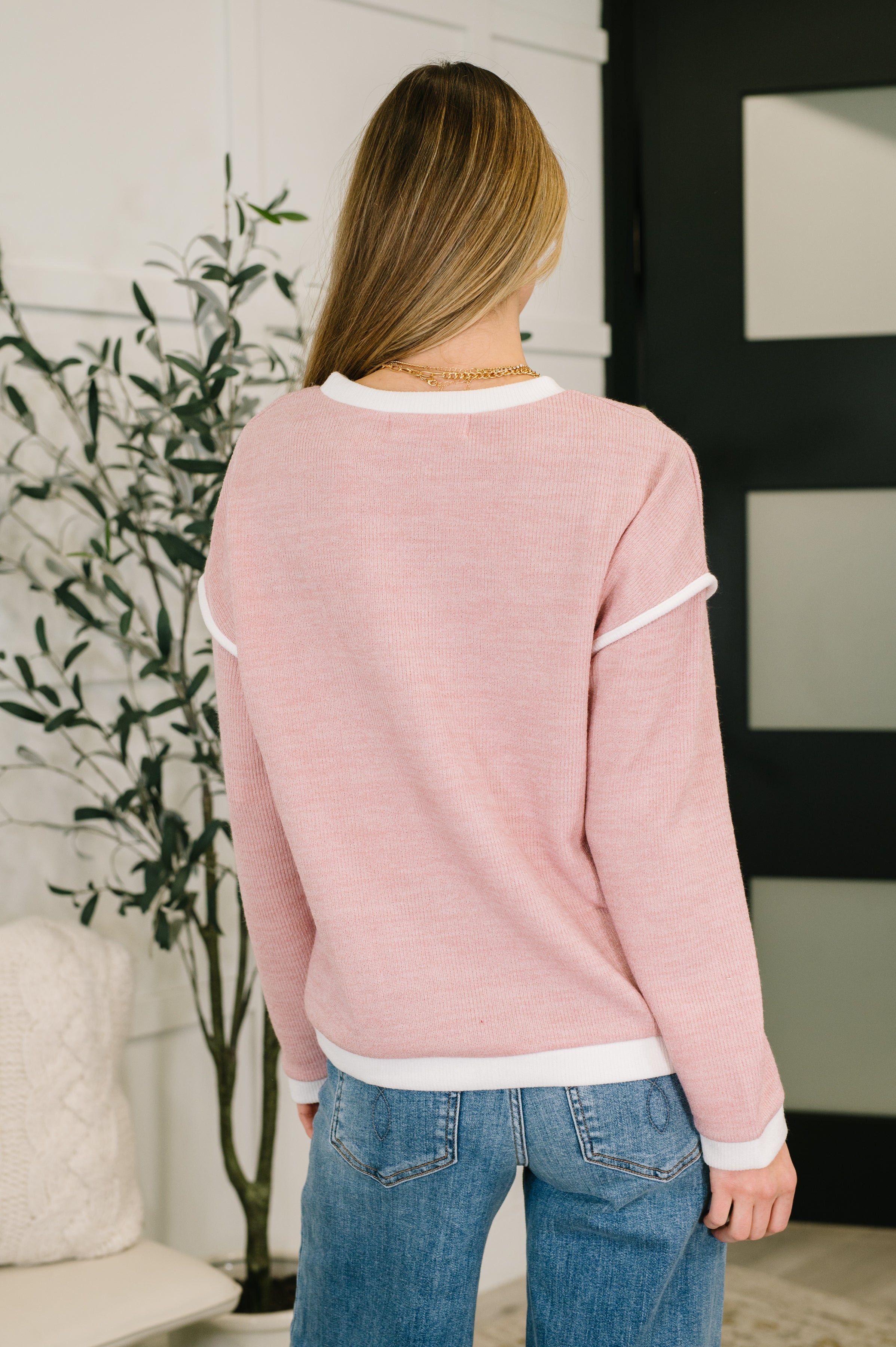 Back view of woman wearing a soft pink textured drop shoulder top with white trim and blue jeans indoors