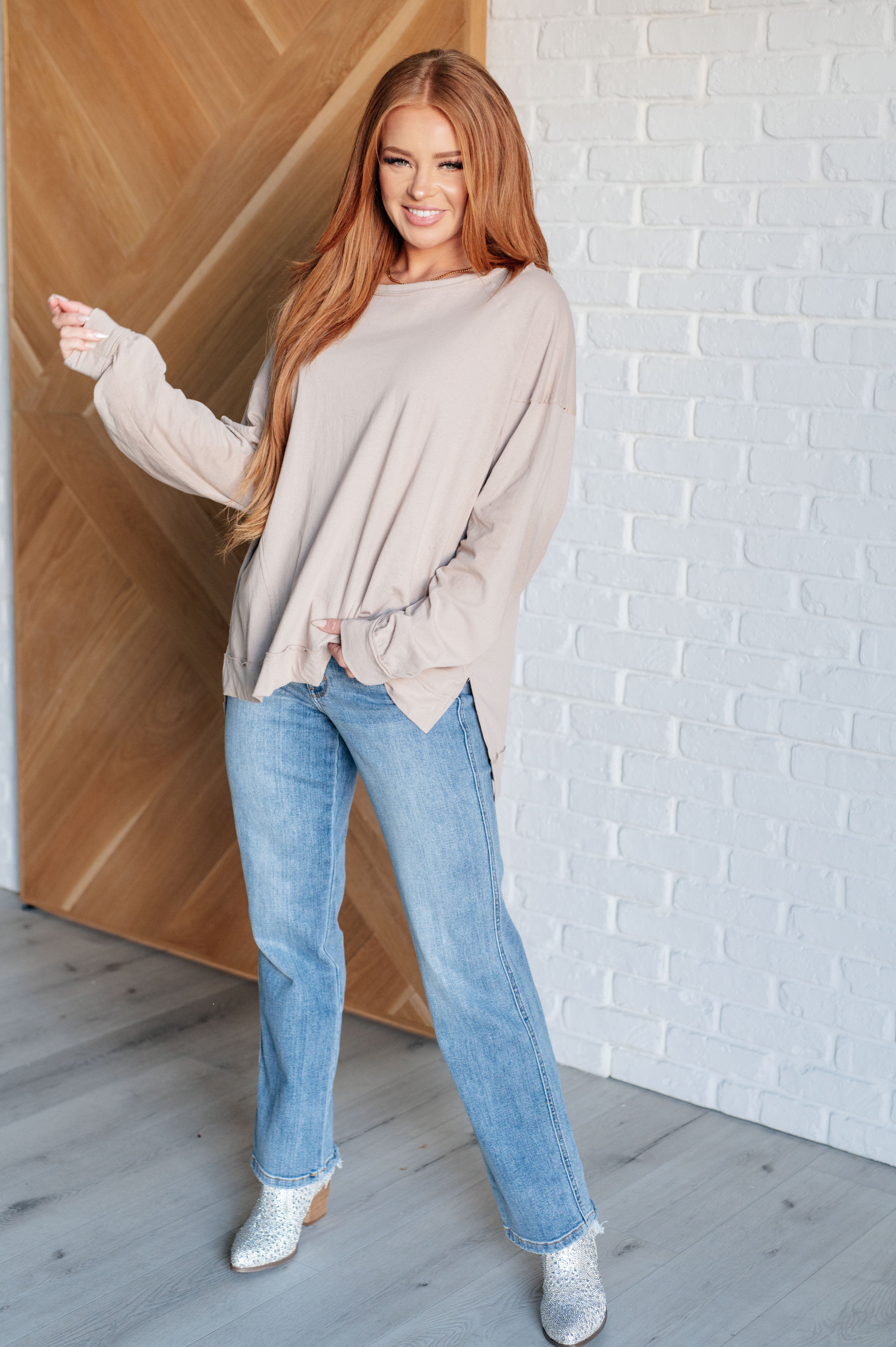 Smiling woman with long red hair wearing oversized beige long sleeve top and light blue jeans indoors