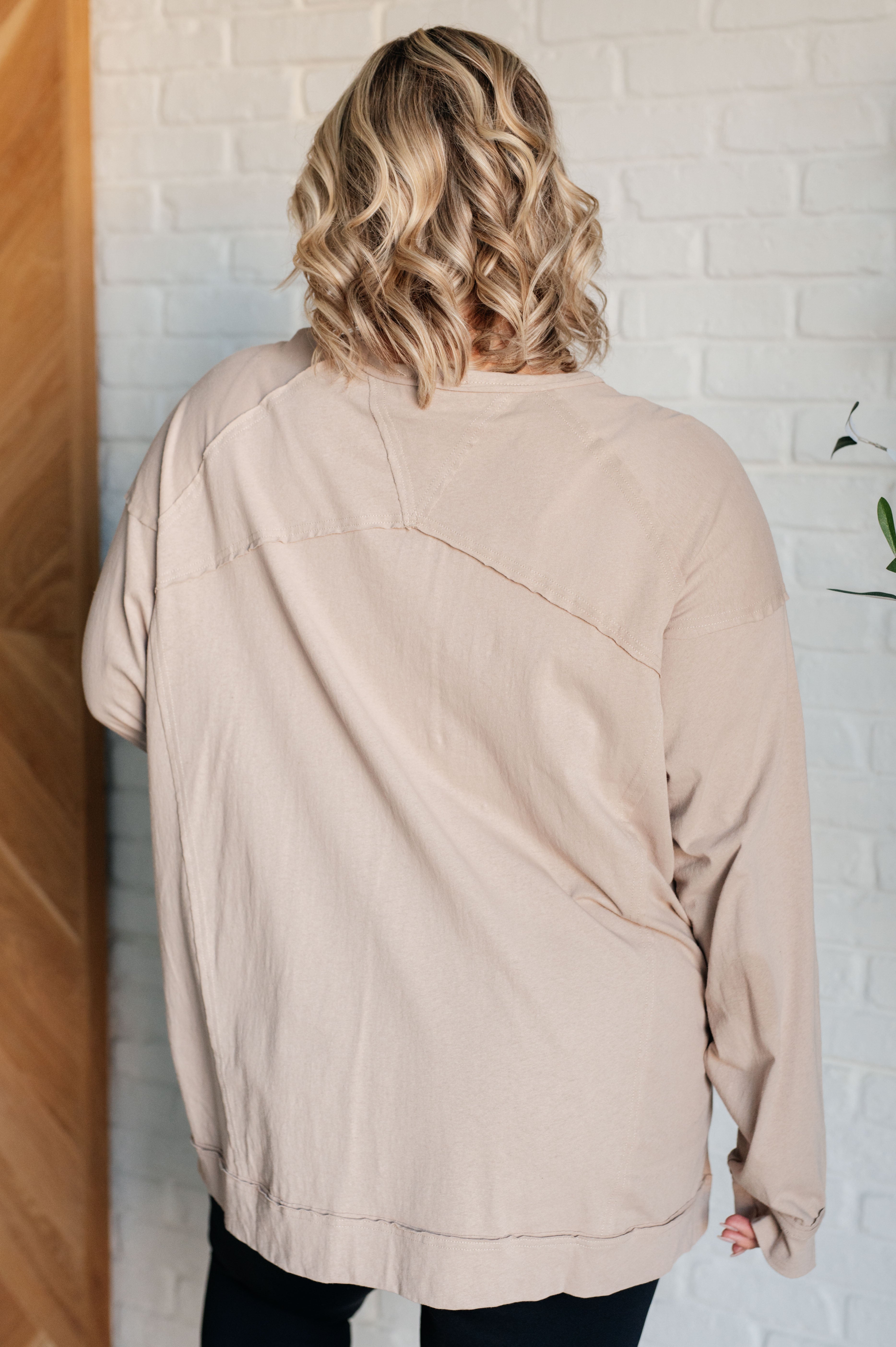 Rear view of person wearing a beige long sleeve top with visible seam details indoors against white brick wall