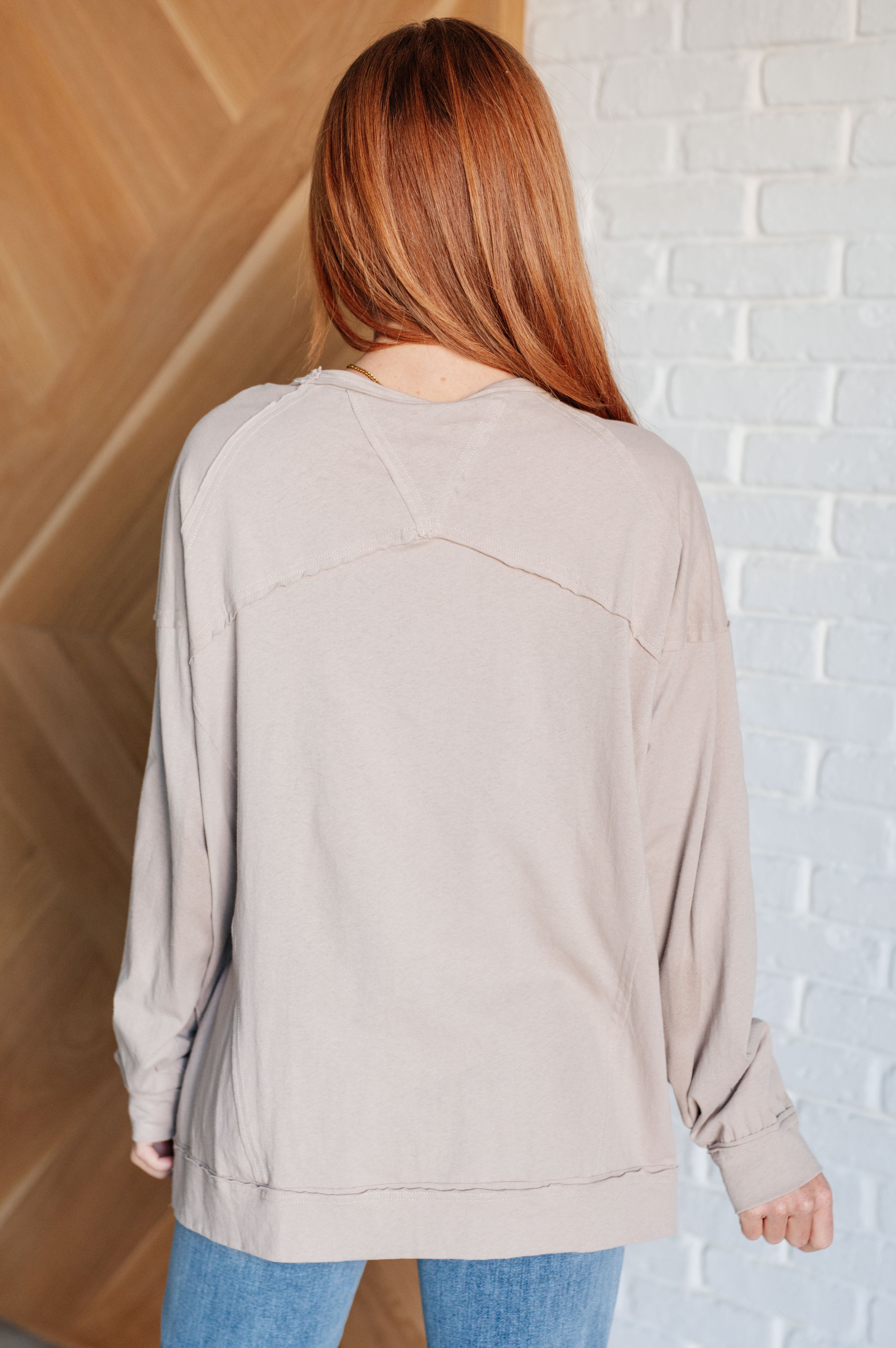 Back view of woman wearing beige long sleeve top with seam details and blue jeans indoors against white brick wall