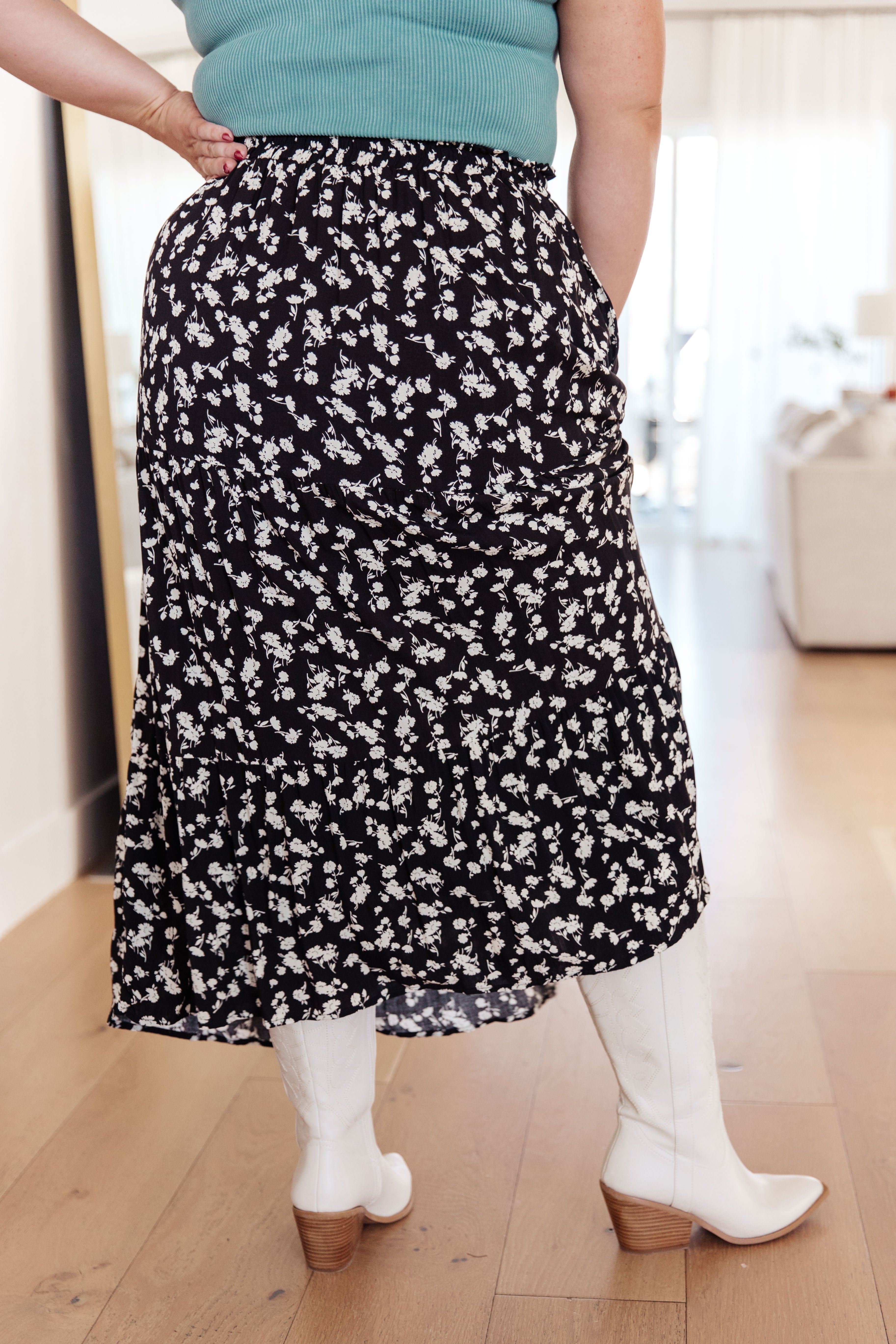 Back view of a woman wearing a black floral skirt with side pockets and white mid-calf boots indoors