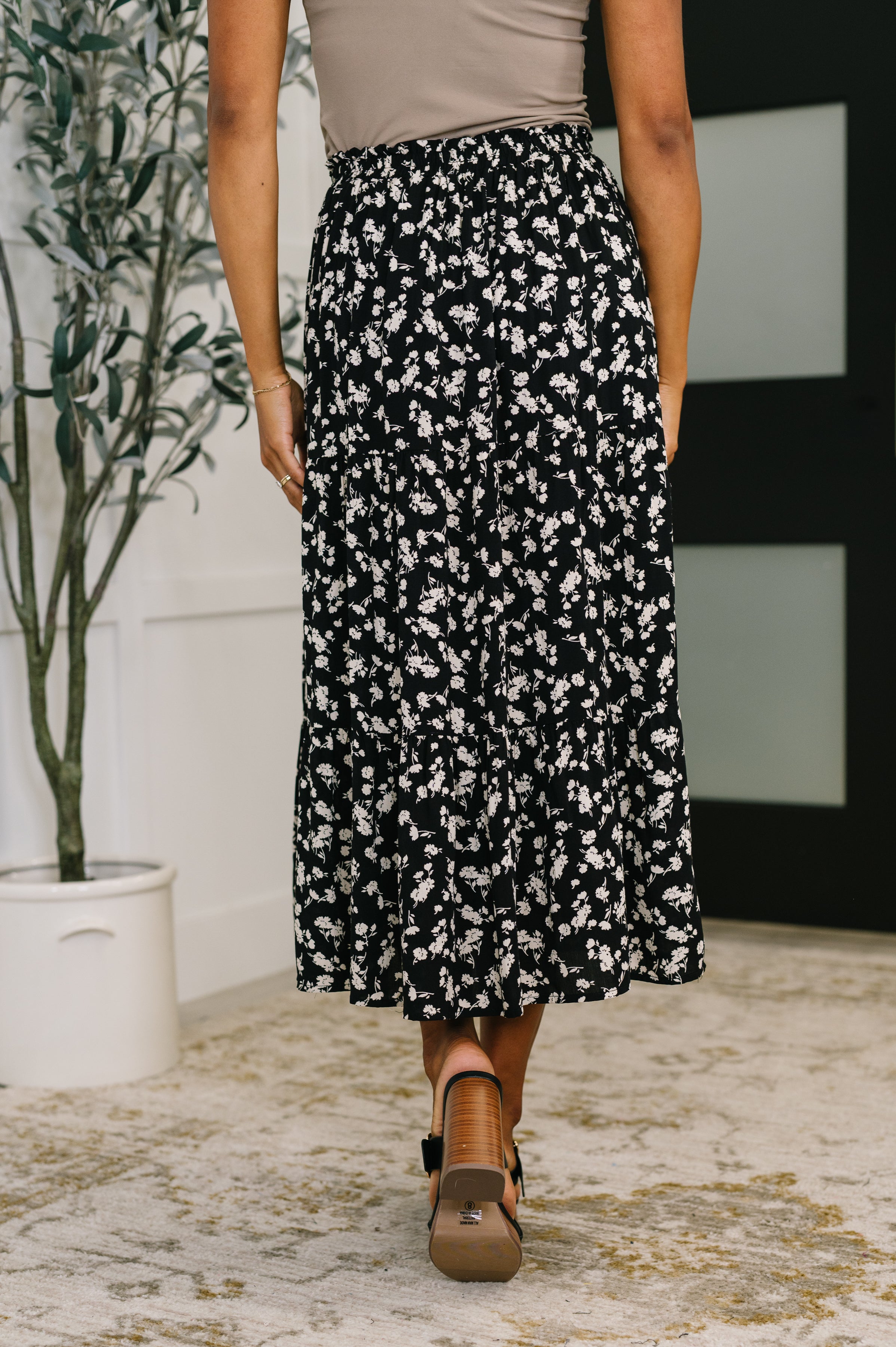 Back view of a woman wearing a flowing black floral skirt with a fitted waist and tan block-heeled sandals indoors