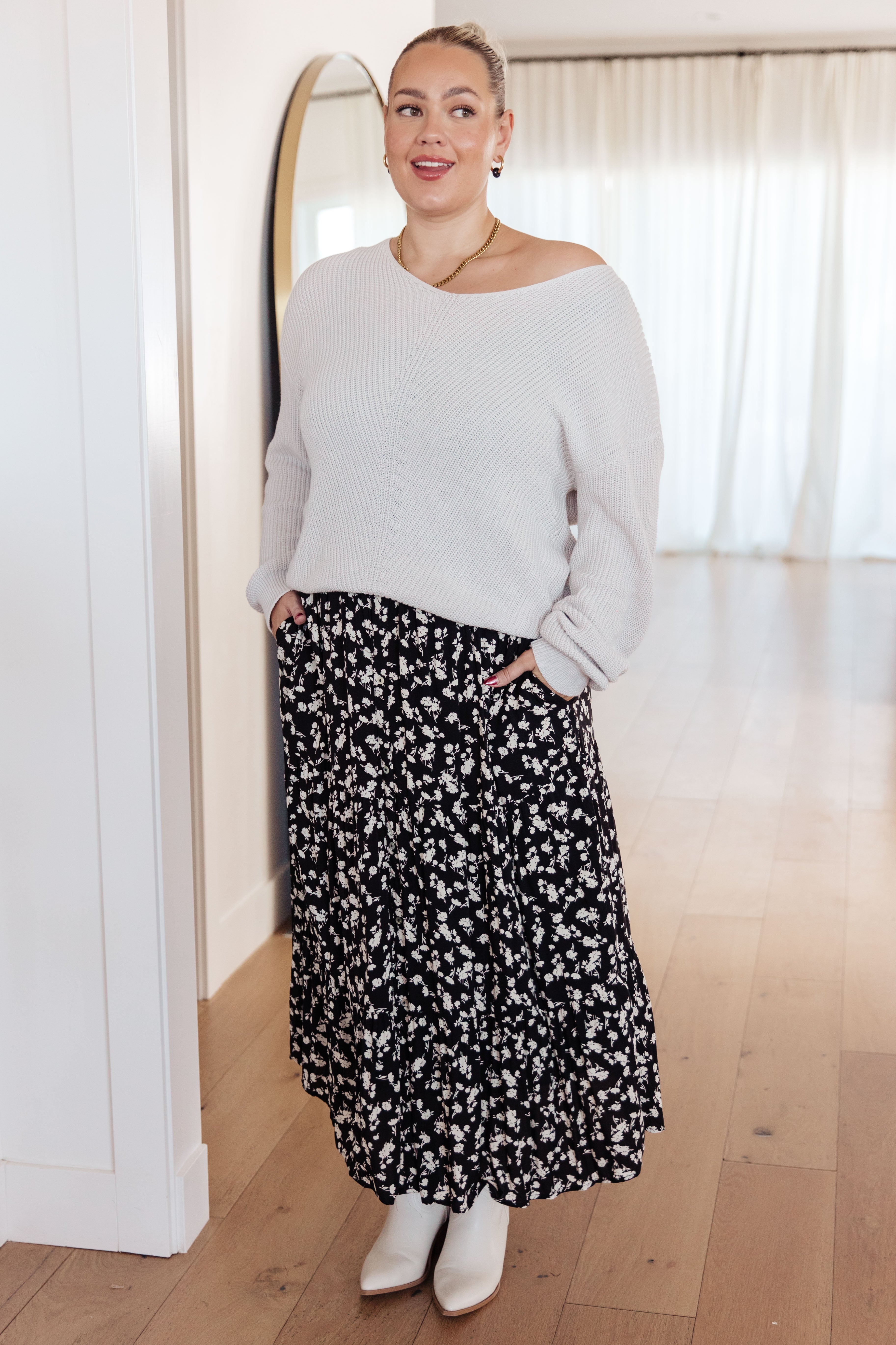 Woman wearing a black ditsy floral skirt with side pockets, paired with a cream knit sweater and white ankle boots indoors
