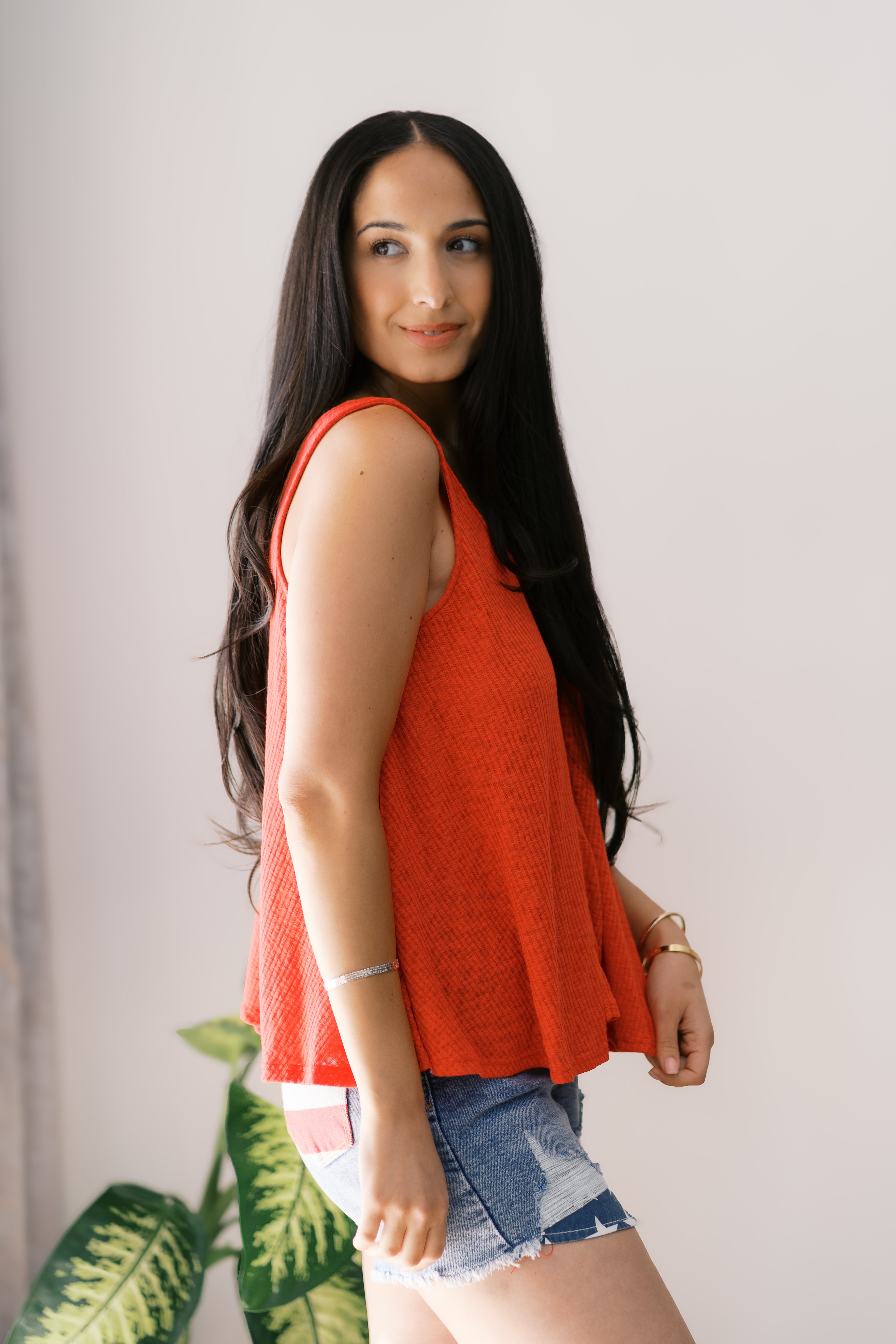 woman with long dark hair wearing a loose bright orange sleeveless tank top and ripped denim shorts indoors