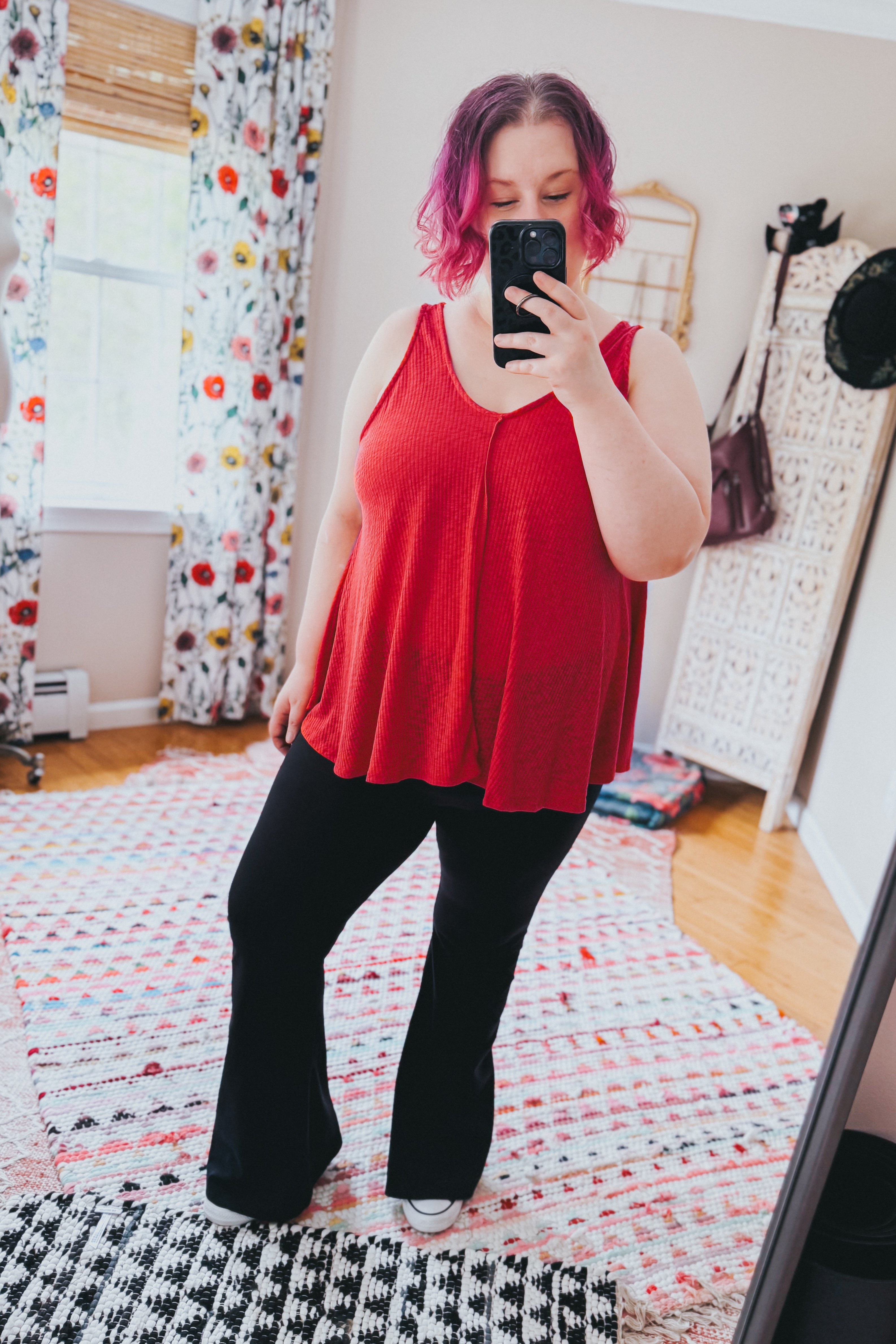 Plus size woman with pink hair wearing a loose red sleeveless tank top and black pants indoors taking a mirror selfie