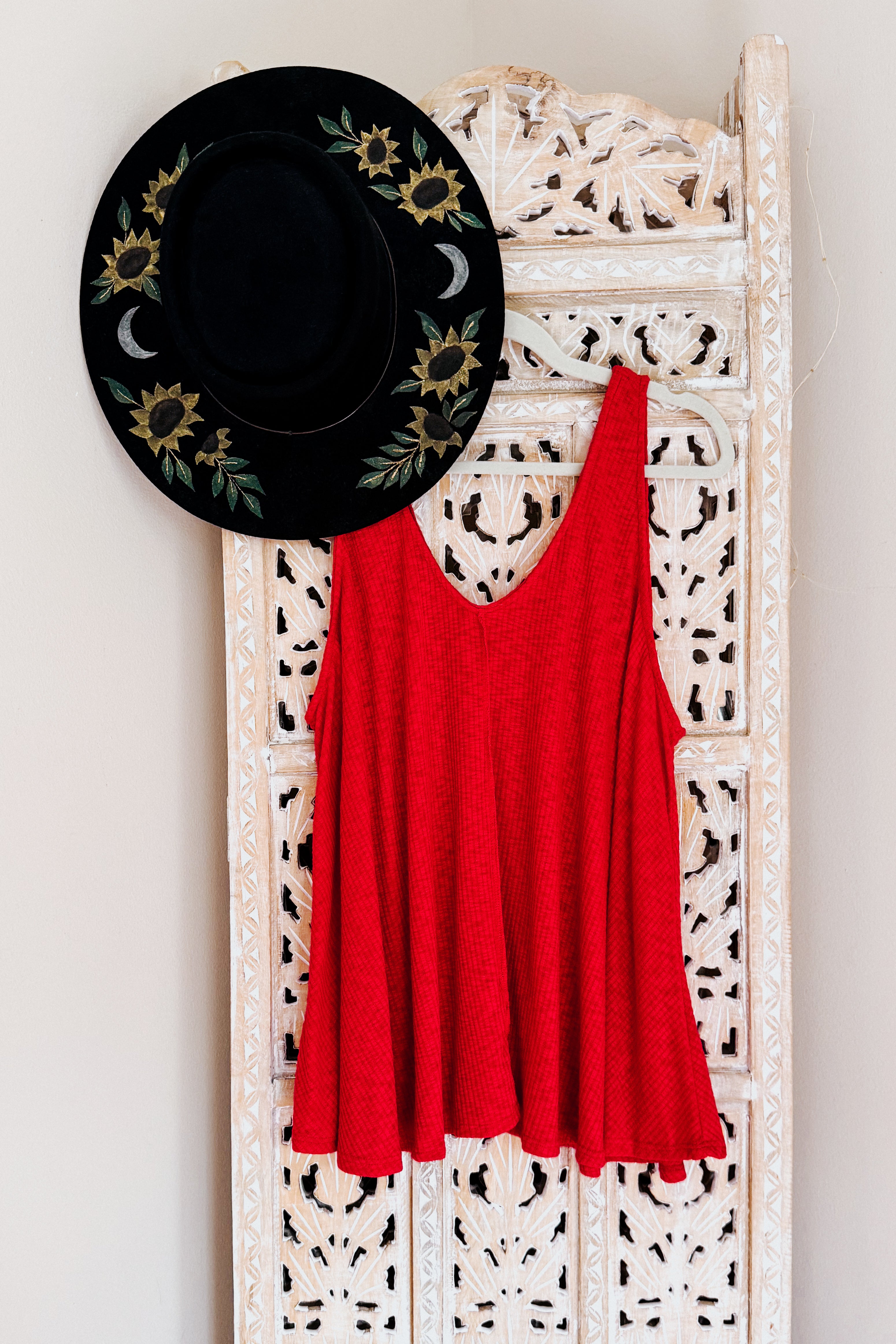 Red textured sleeveless tank top hanging on ornate white wooden screen with black hat decorated with sunflowers and moons