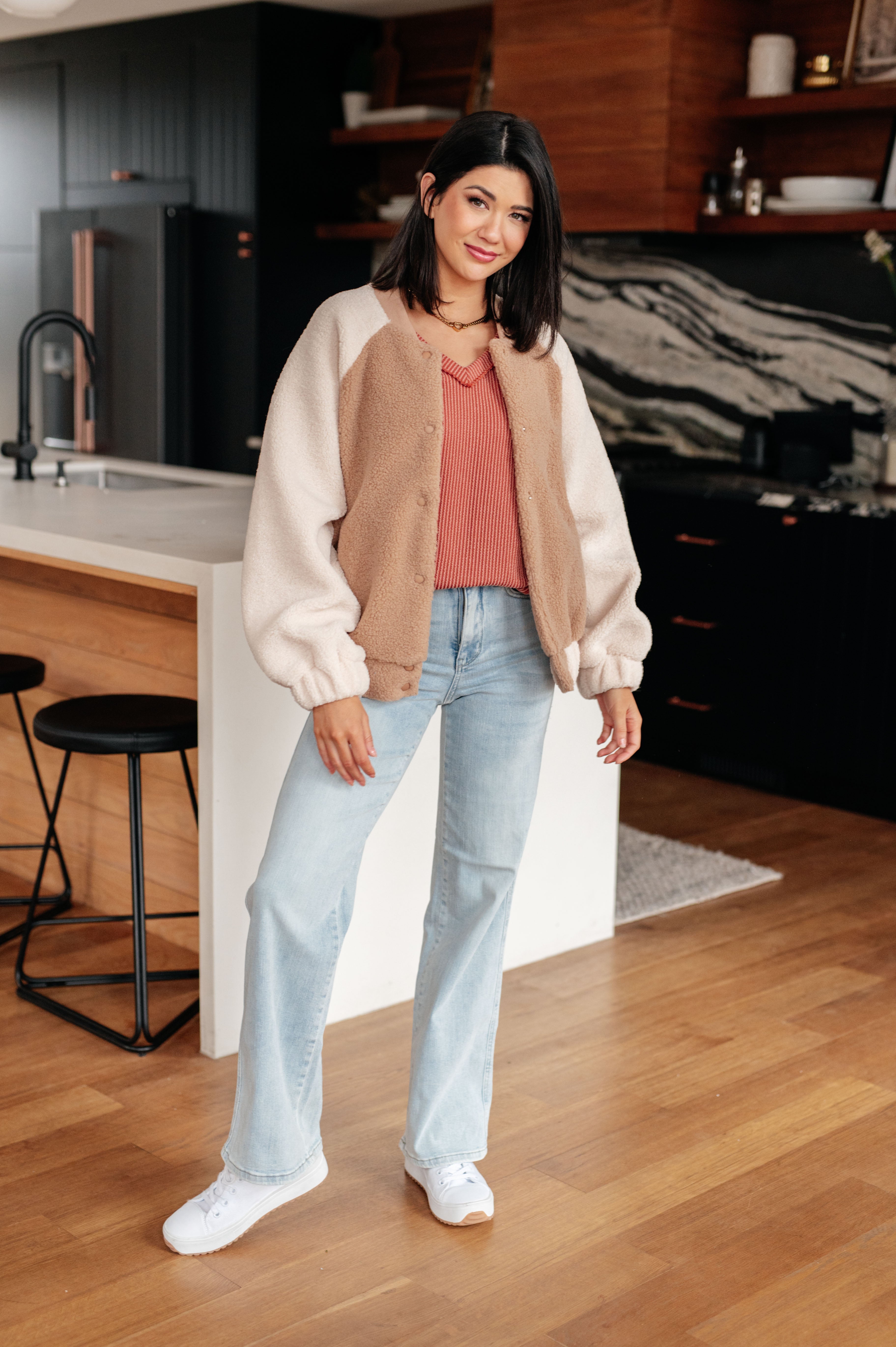 Woman smiling indoors wearing rust First and Foremost Rib Knit Top styled with light wash jeans and white sneakers in a modern kitchen environment