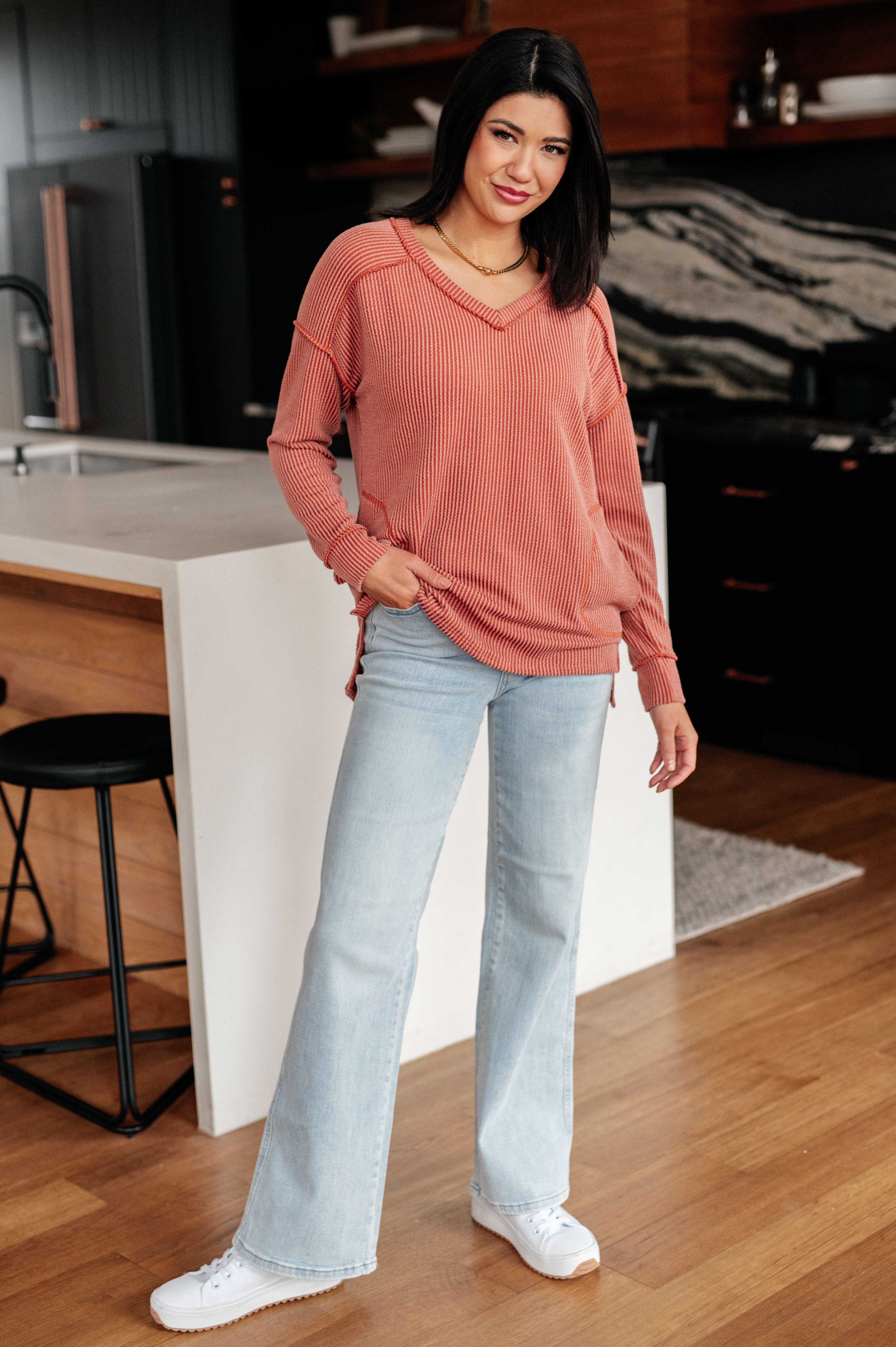 Woman posing indoors wearing rust First and Foremost Rib Knit Top with V-neck and relaxed fit paired with light wash flared jeans and white sneakers in modern kitchen