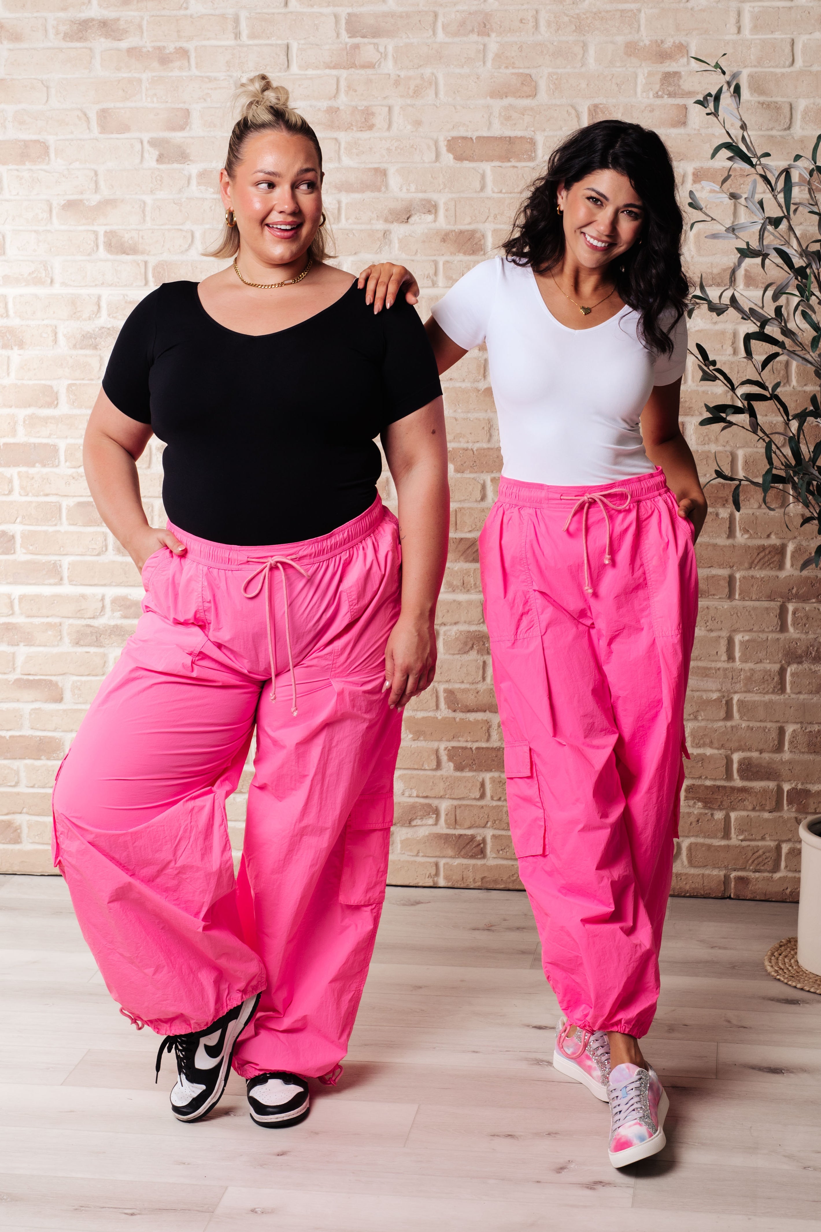 Two women posing indoors wearing bright fuchsia First Place Cargo Pants styled with casual black and white tops and sneakers against a brick wall background