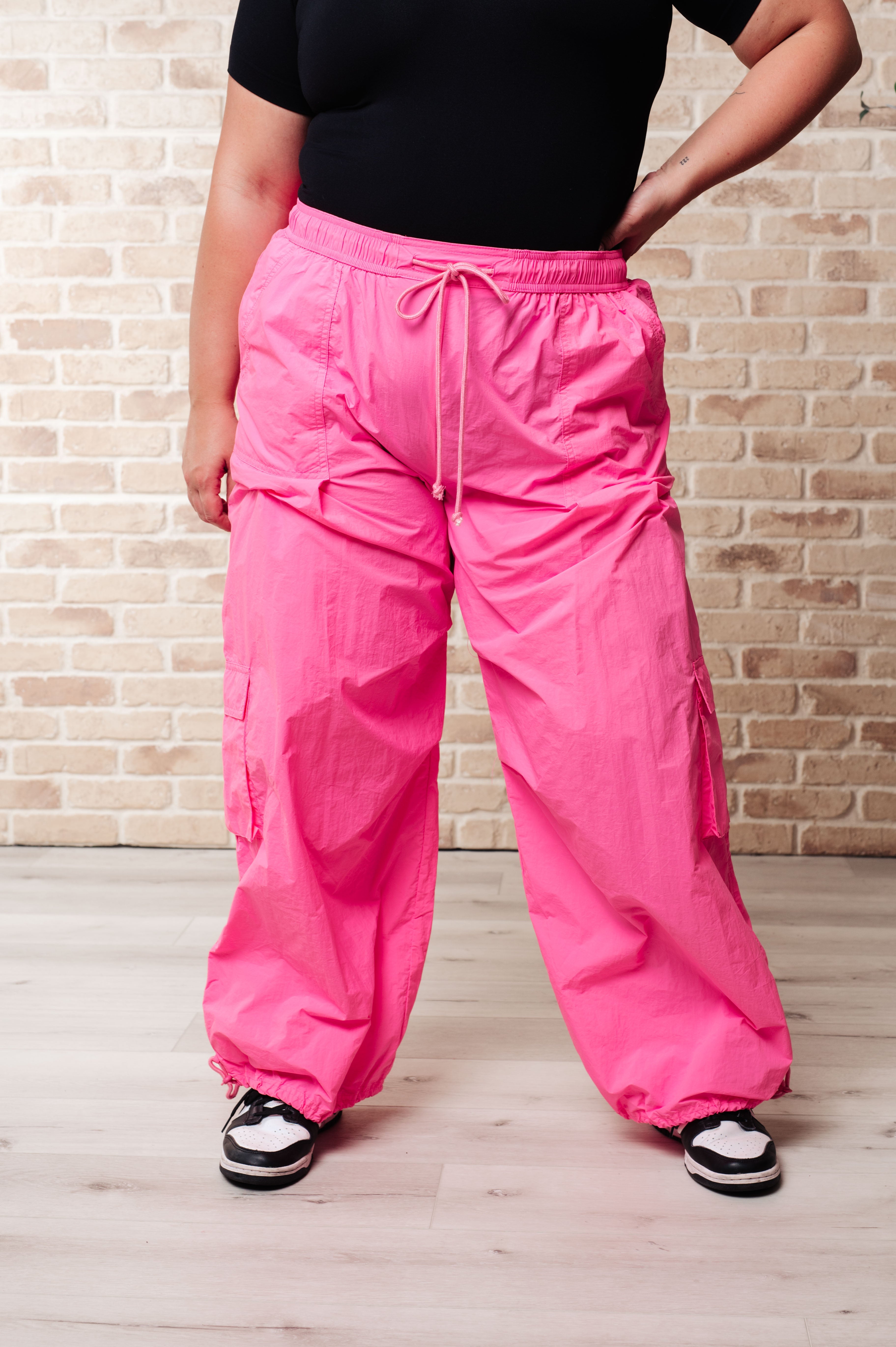 Front view of woman wearing bright fuchsia First Place Cargo Pants with drawstring waist and elastic cuffs styled with sneakers against a brick wall background