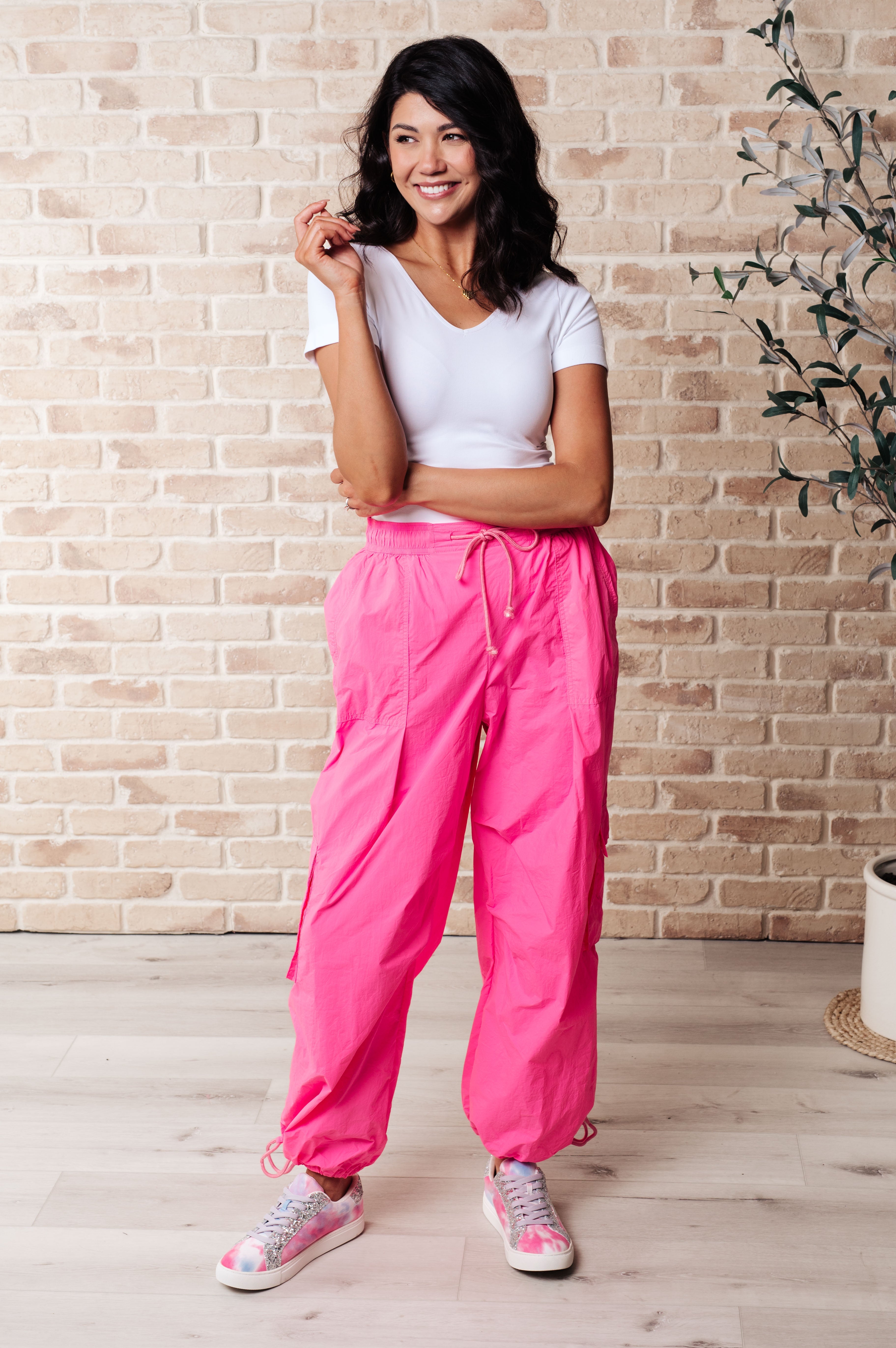 Smiling woman wearing bright fuchsia First Place Cargo Pants with drawstring waist and ankle ties paired with casual white T-shirt and pink sneakers indoors