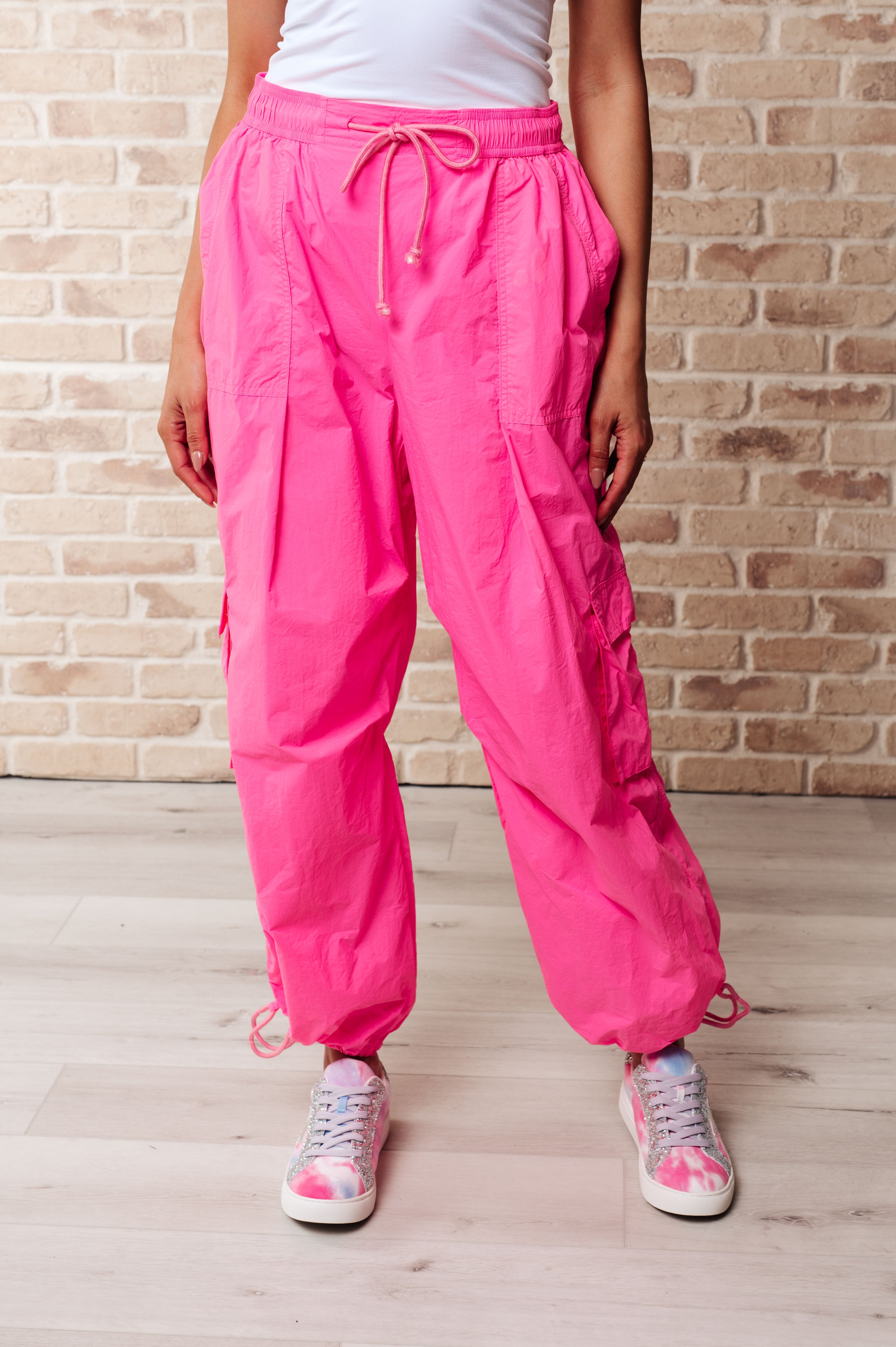 Front view of woman wearing bright fuchsia First Place Cargo Pants with drawstring waist and ankle ties paired with white sneakers on light wooden floor