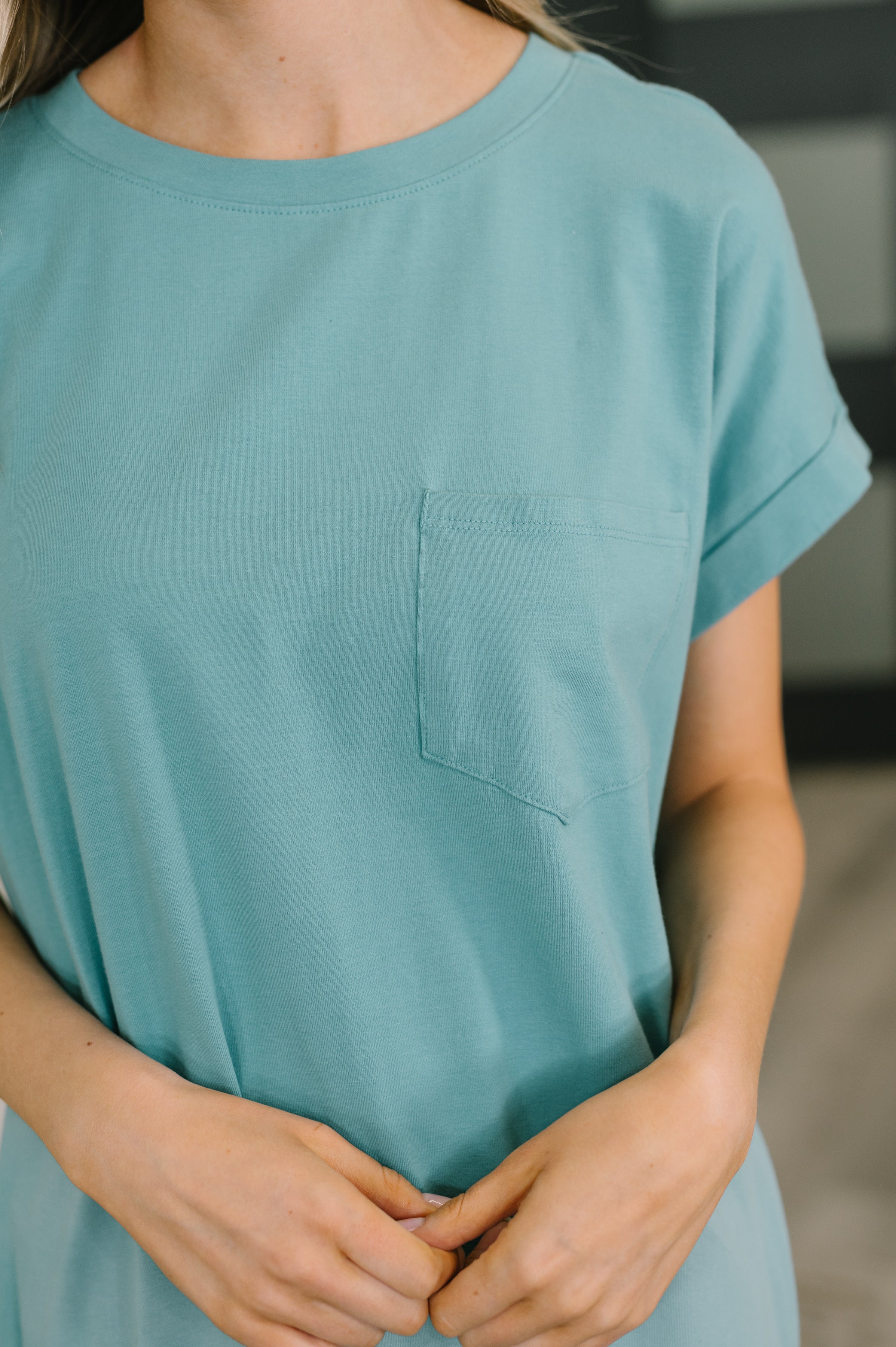 Close-up of woman wearing dusty teal short sleeve midi dress with ribbed round neckline and chest pocket indoors
