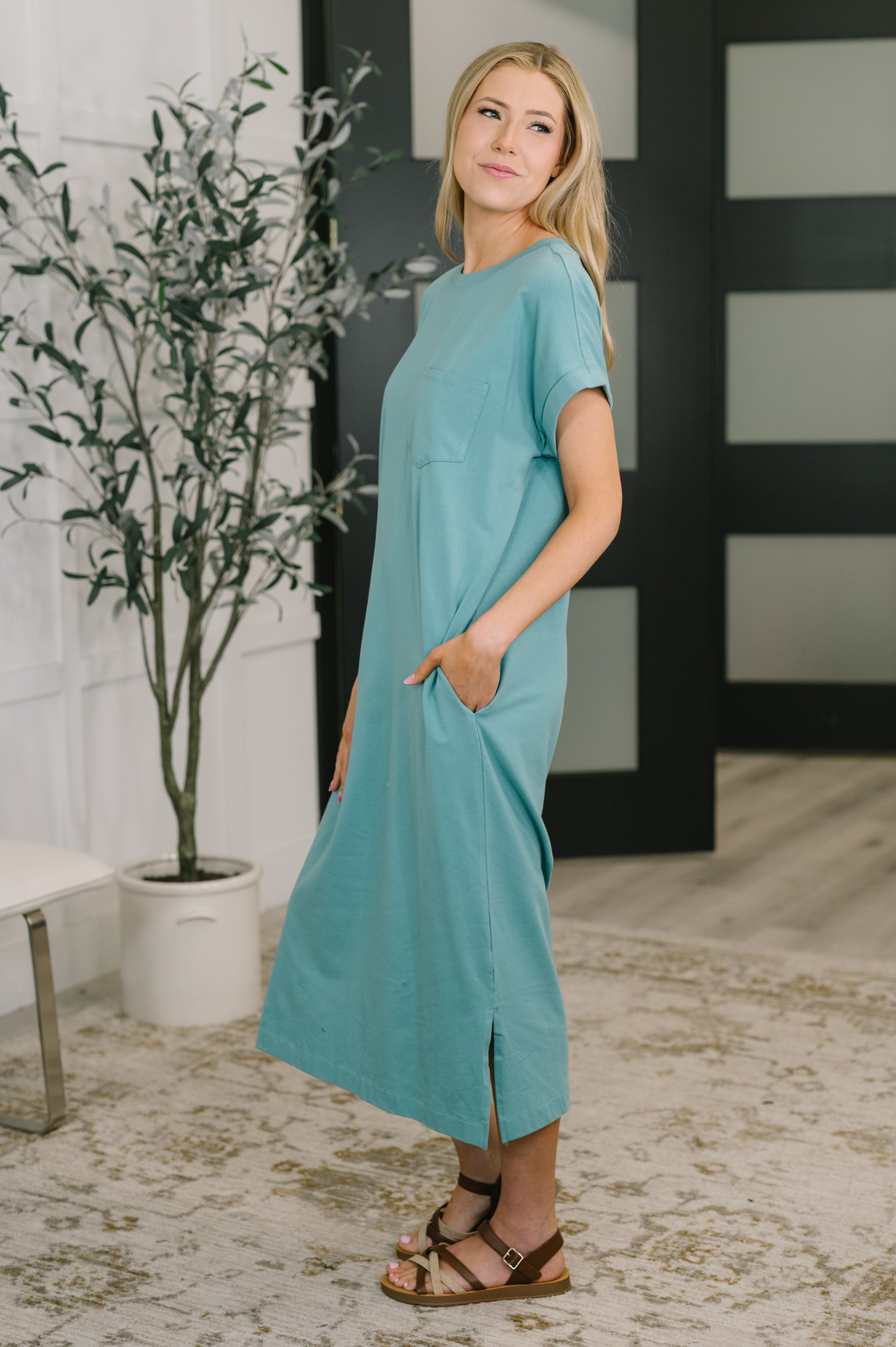 Side profile of woman in dusty teal short sleeve midi dress with pockets and brown sandals indoors
