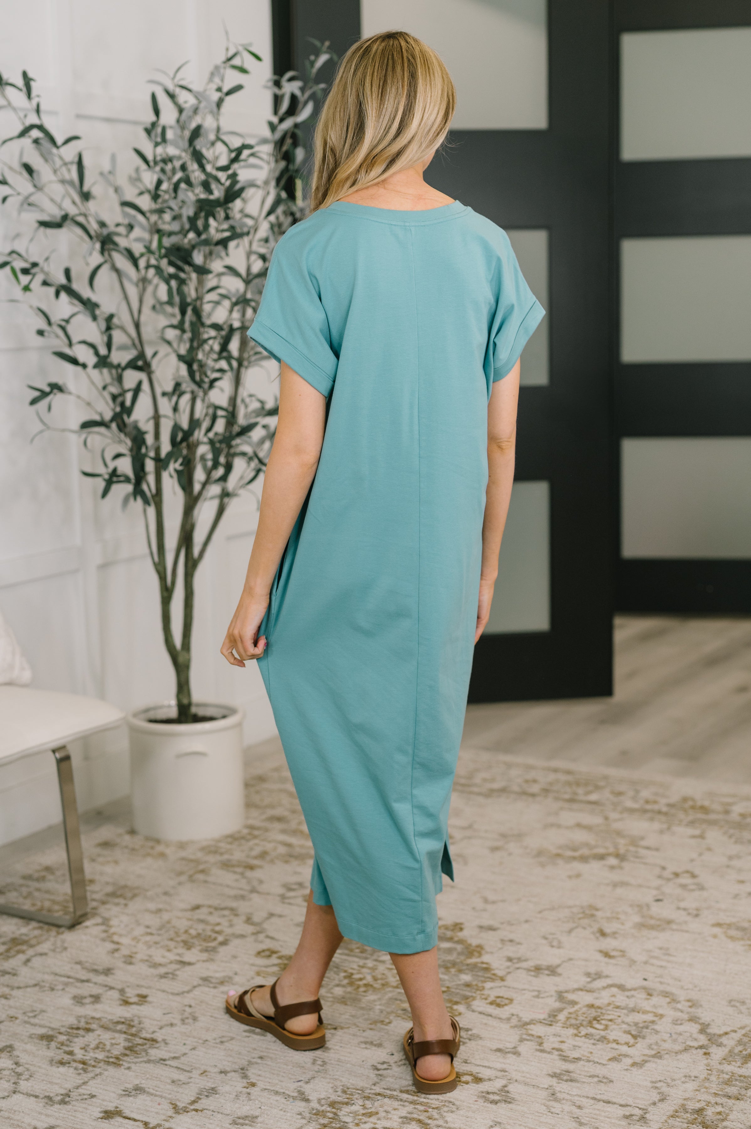 Rear view of woman wearing dusty teal midi dress with short sleeves and side slits paired with brown sandals indoors