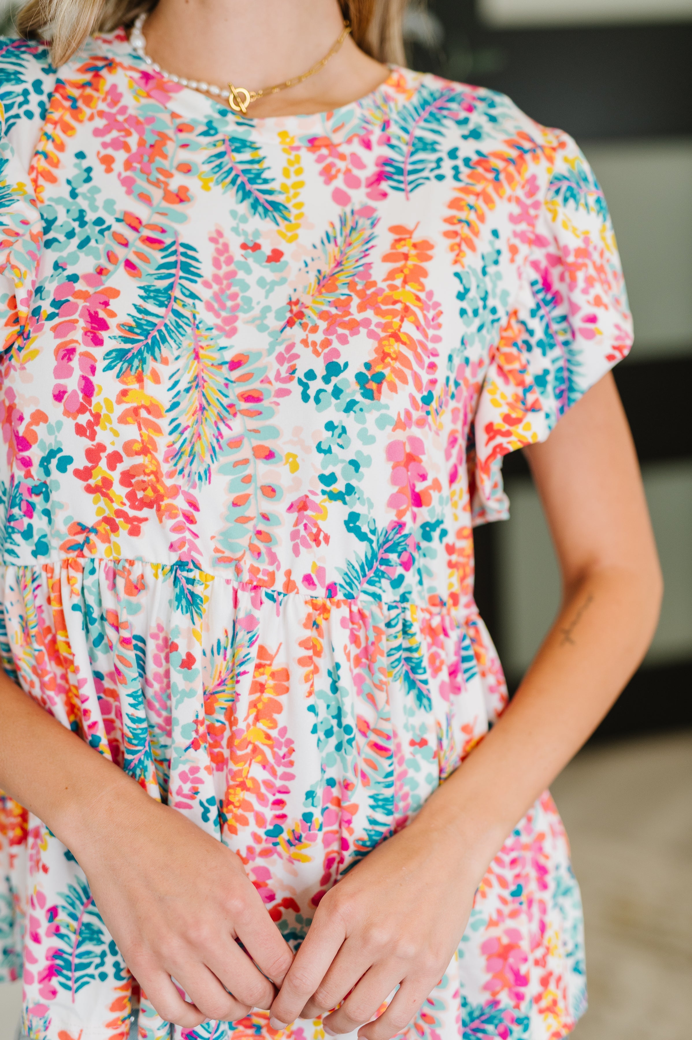 Flirt Alert Flutter Sleeve Top • Ivory with bright floral and leaf pattern, flutter sleeves, and gathered waist worn by woman captured from the shoulders to waist.