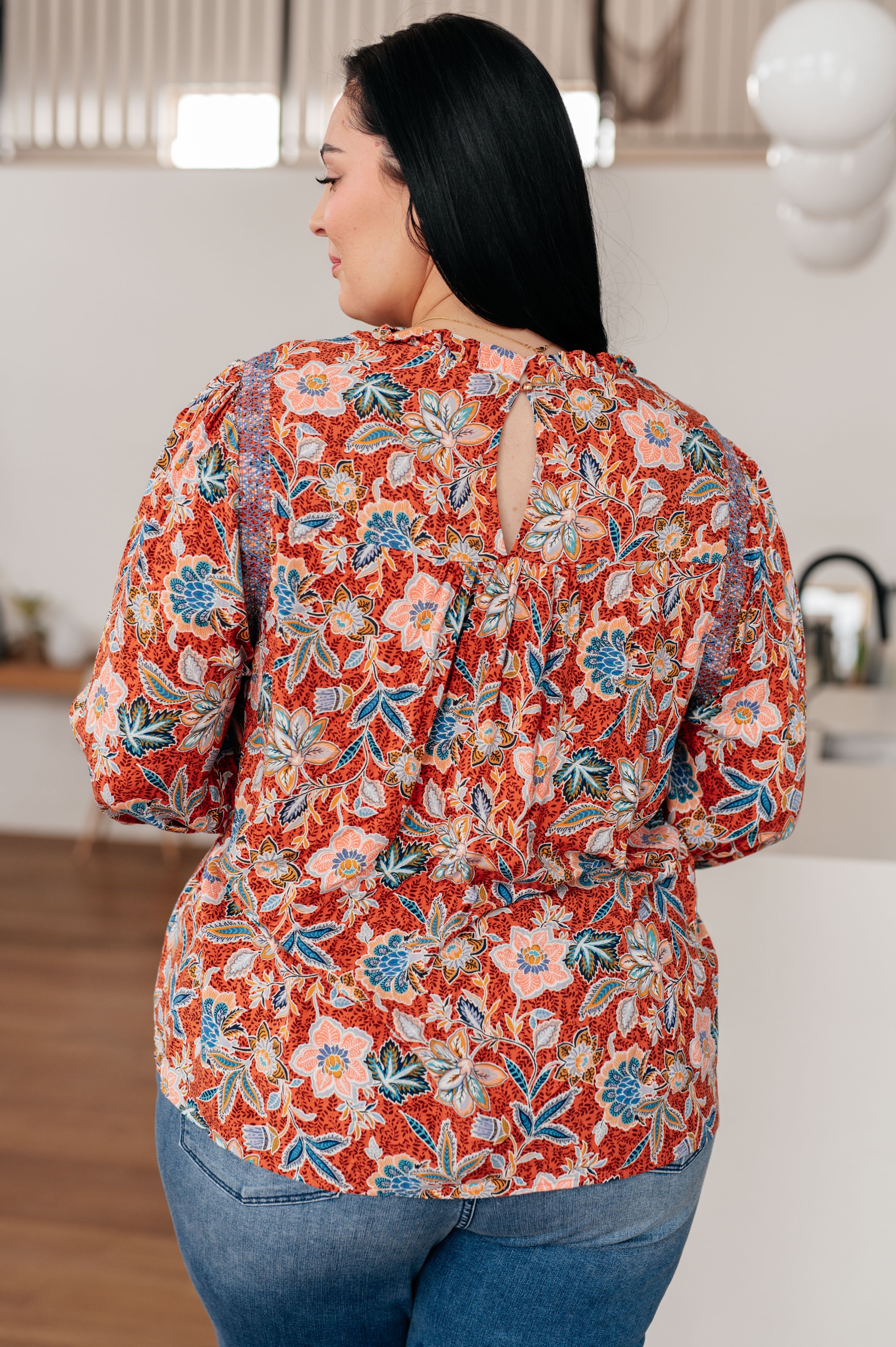 Woman wearing rust floral blouse with keyhole back detail paired with blue jeans in cozy indoor setting