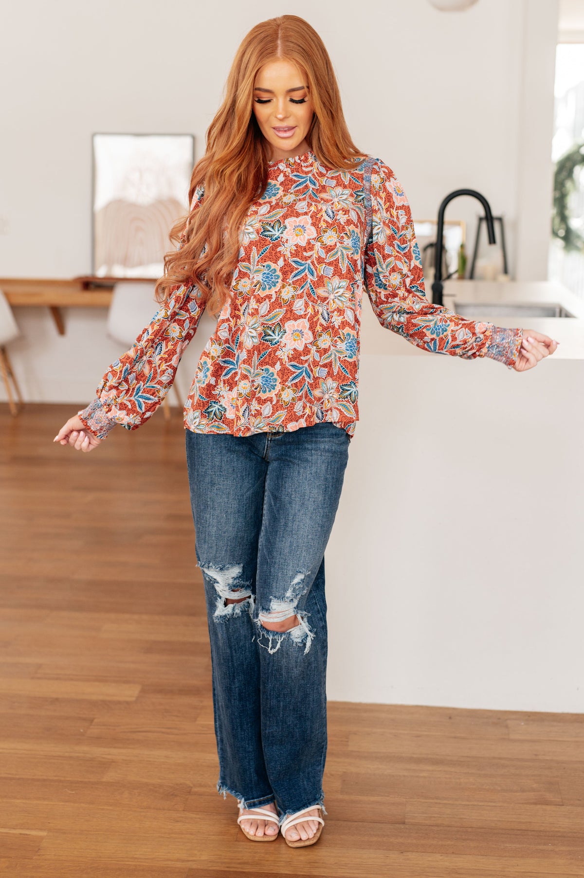 Woman modeling a rust floral blouse with a ruffled neckline and long sleeves, distressed jeans, and sandals in a bright home interior