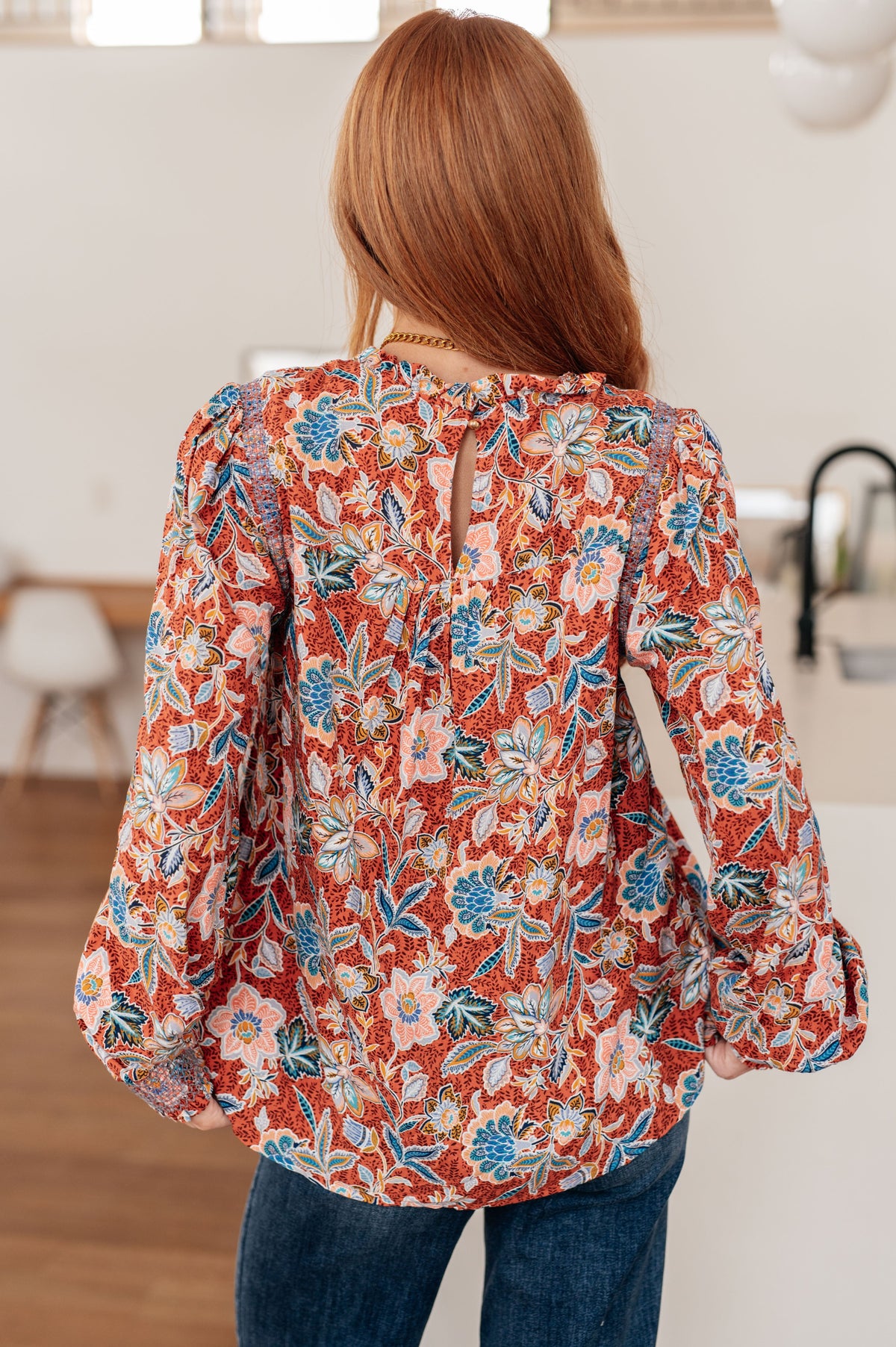 Back view of a woman wearing a Rust floral blouse with a ruffled neckline and long sleeves with blue jeans indoors