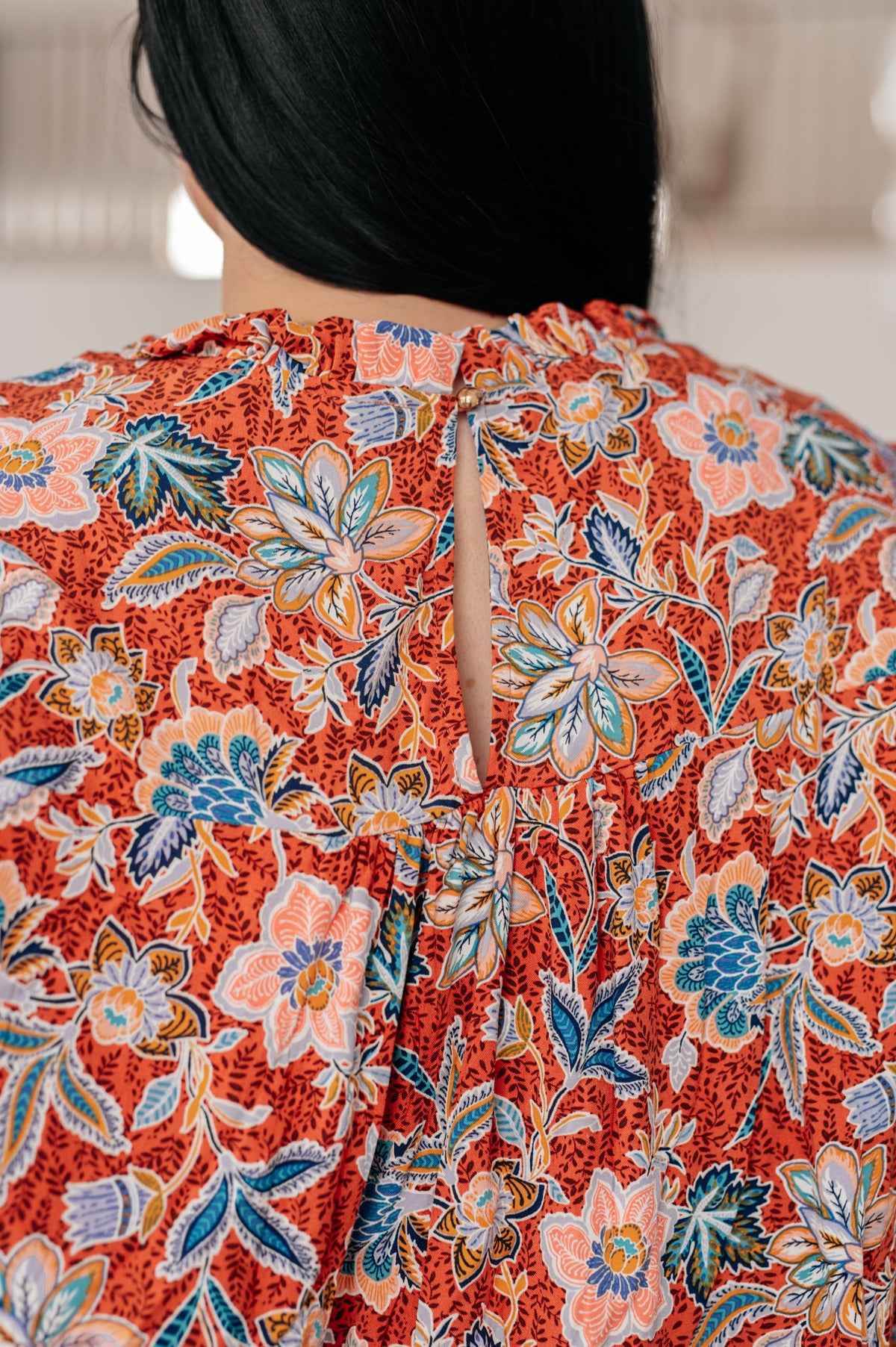 Back view of a woman wearing a rust floral blouse with a ruffled neckline and keyhole back closure indoors