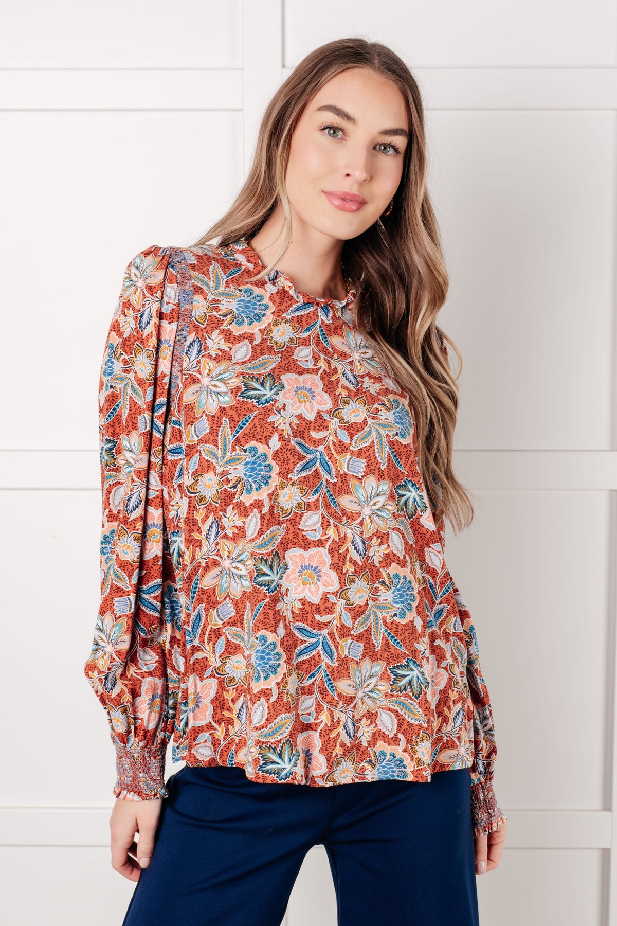Woman modeling a Rust floral blouse with a ruffled neckline and long sleeves paired with navy pants indoors