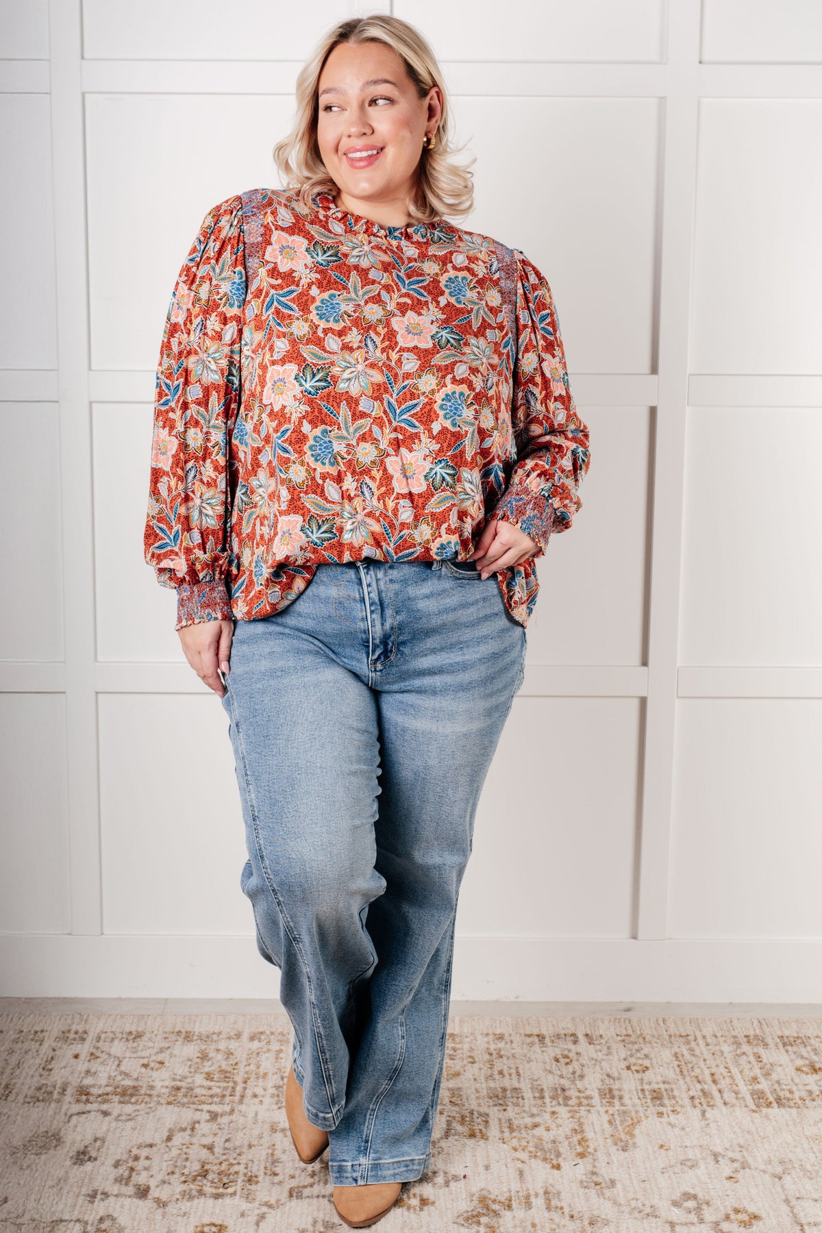 Smiling woman wearing a rust floral blouse with a ruffled neckline and relaxed sleeves paired with light wash jeans indoors