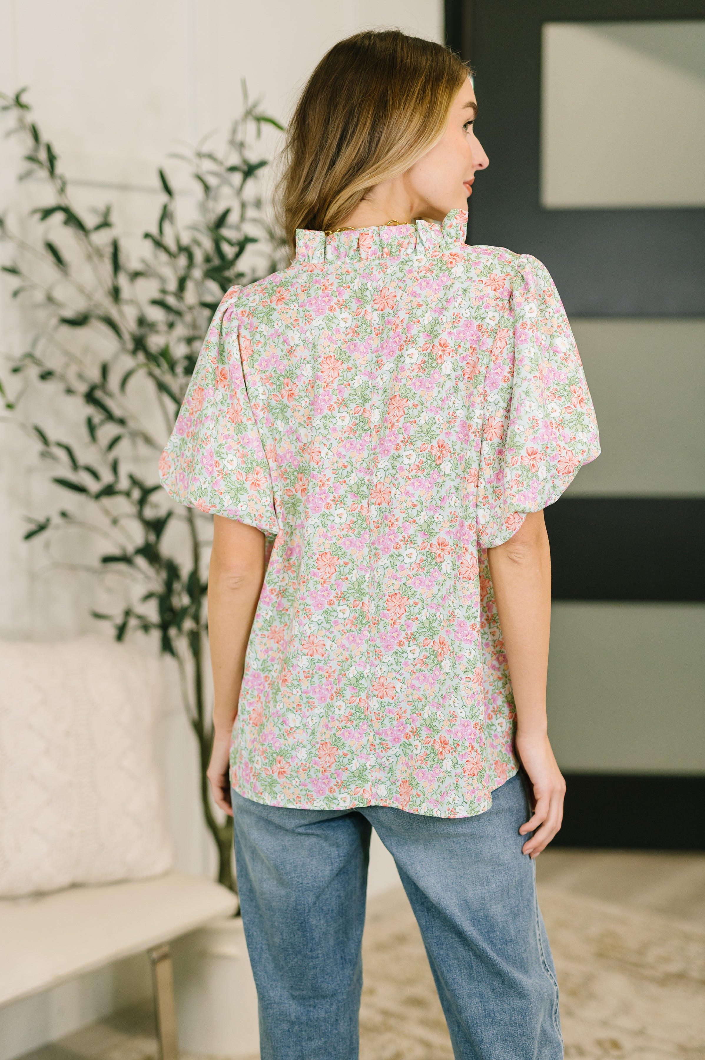 Back view of woman wearing floral blouse with ruffled neckline and voluminous sleeves paired with blue jeans indoors