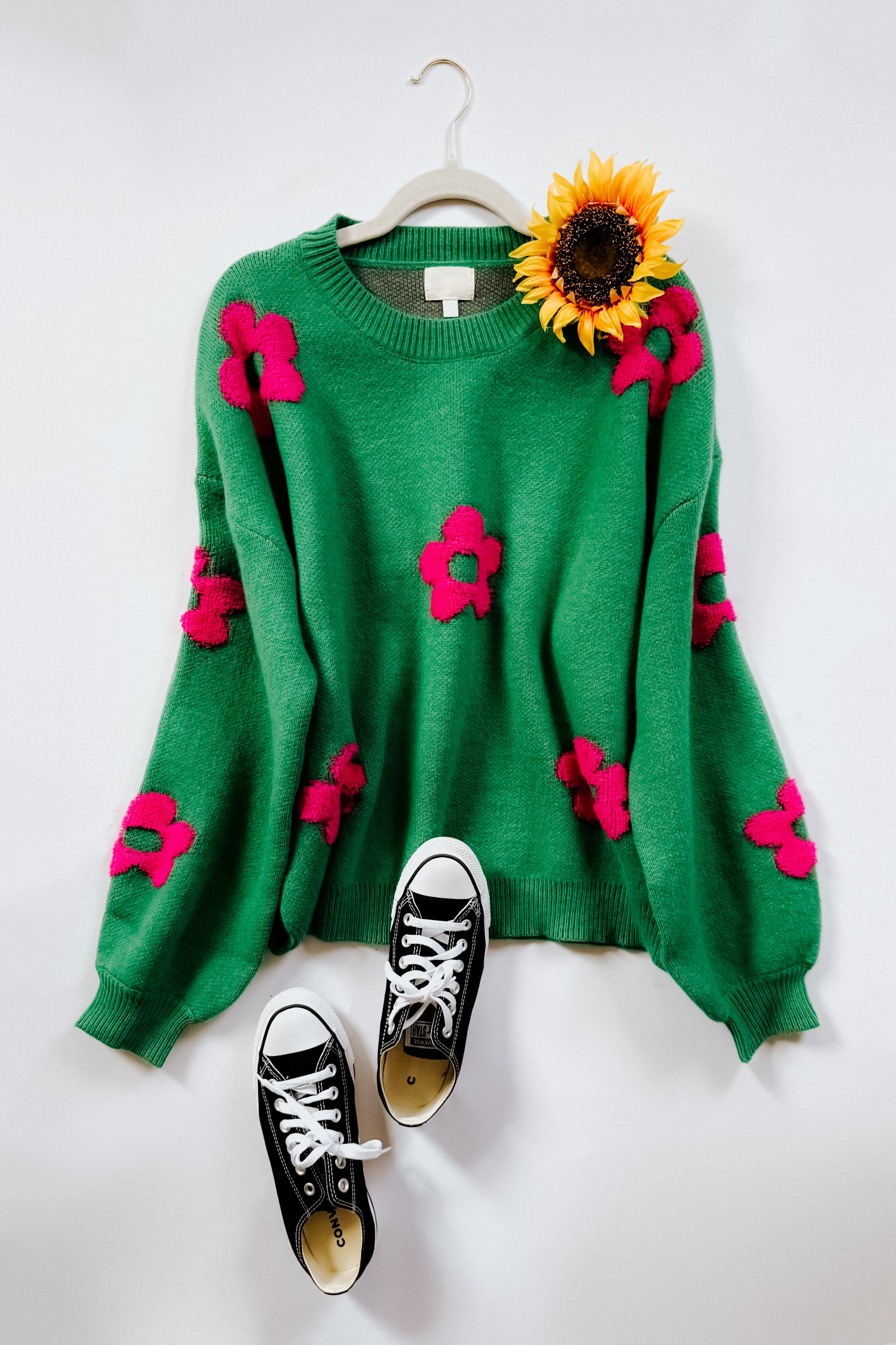 A green sweater with balloon sleeves and a crew neckline, embellished with tufted pink flowers, displayed on a hanger alongside a pair of black converse sneakers on a white flat lay background.