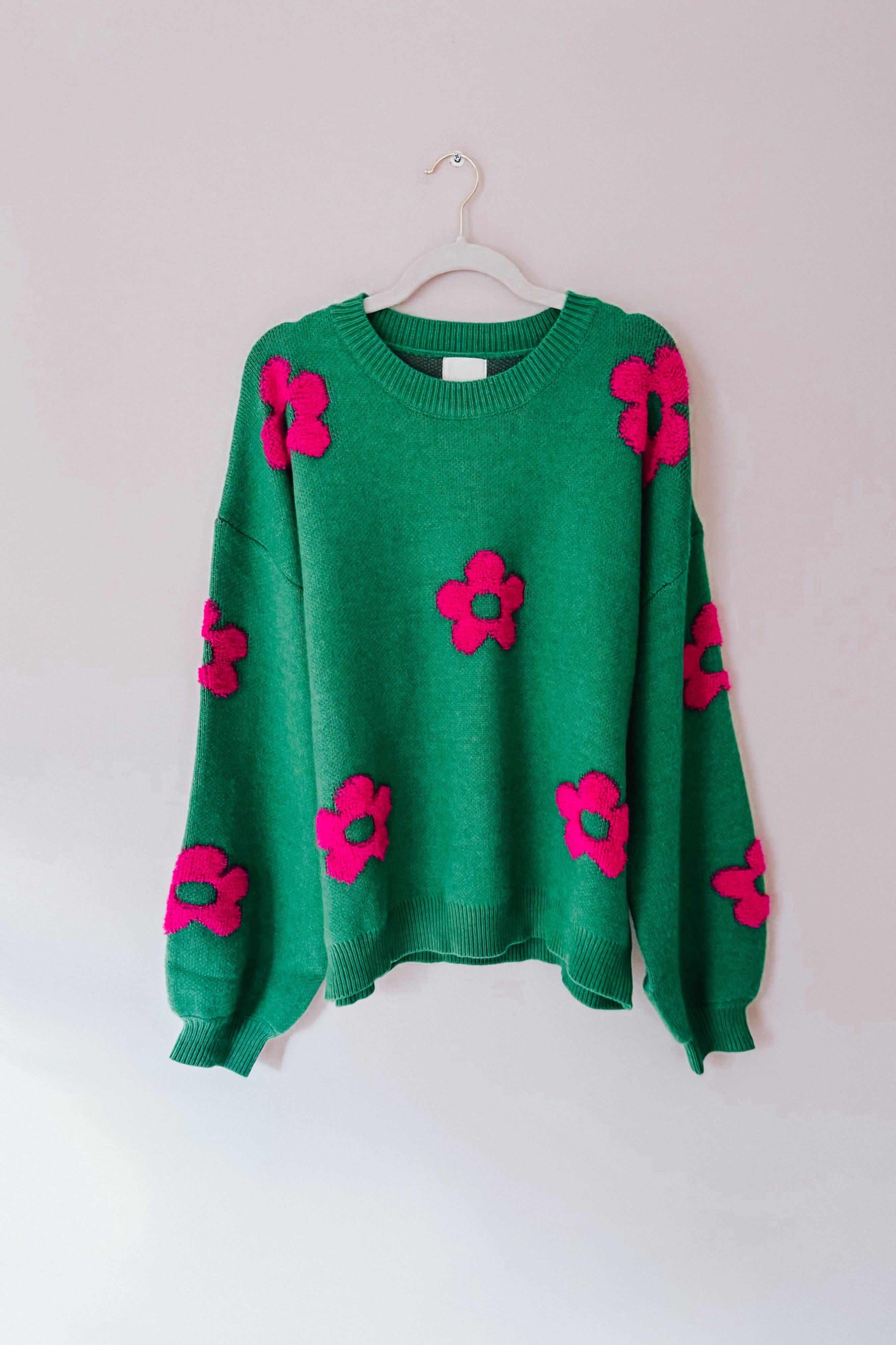 A green sweater embellished with tufted pink flowers hanging on a hanger.