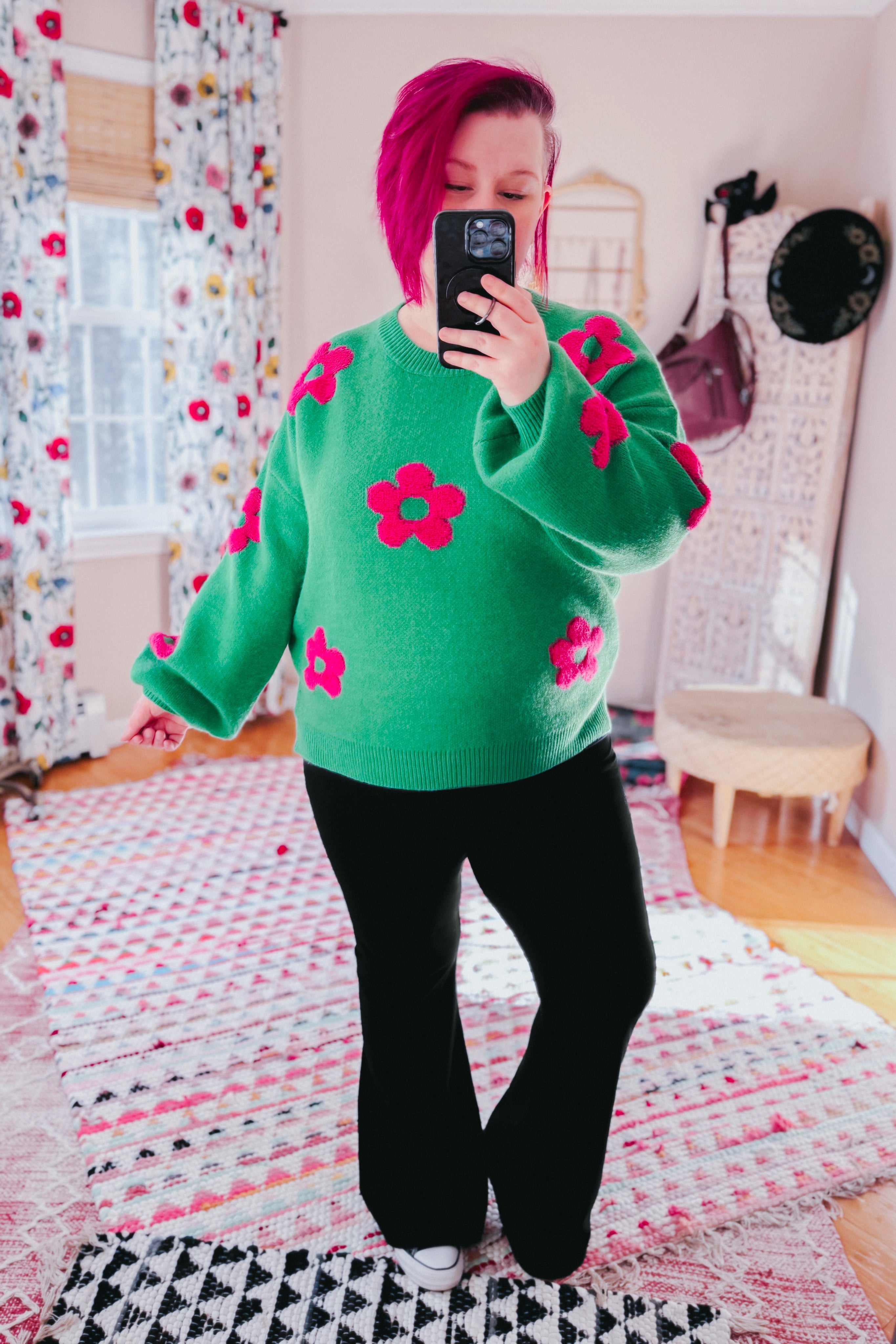 Woman wearing a green sweater embellished with tufted pink flowers and black yoga pants in an indoor setting.