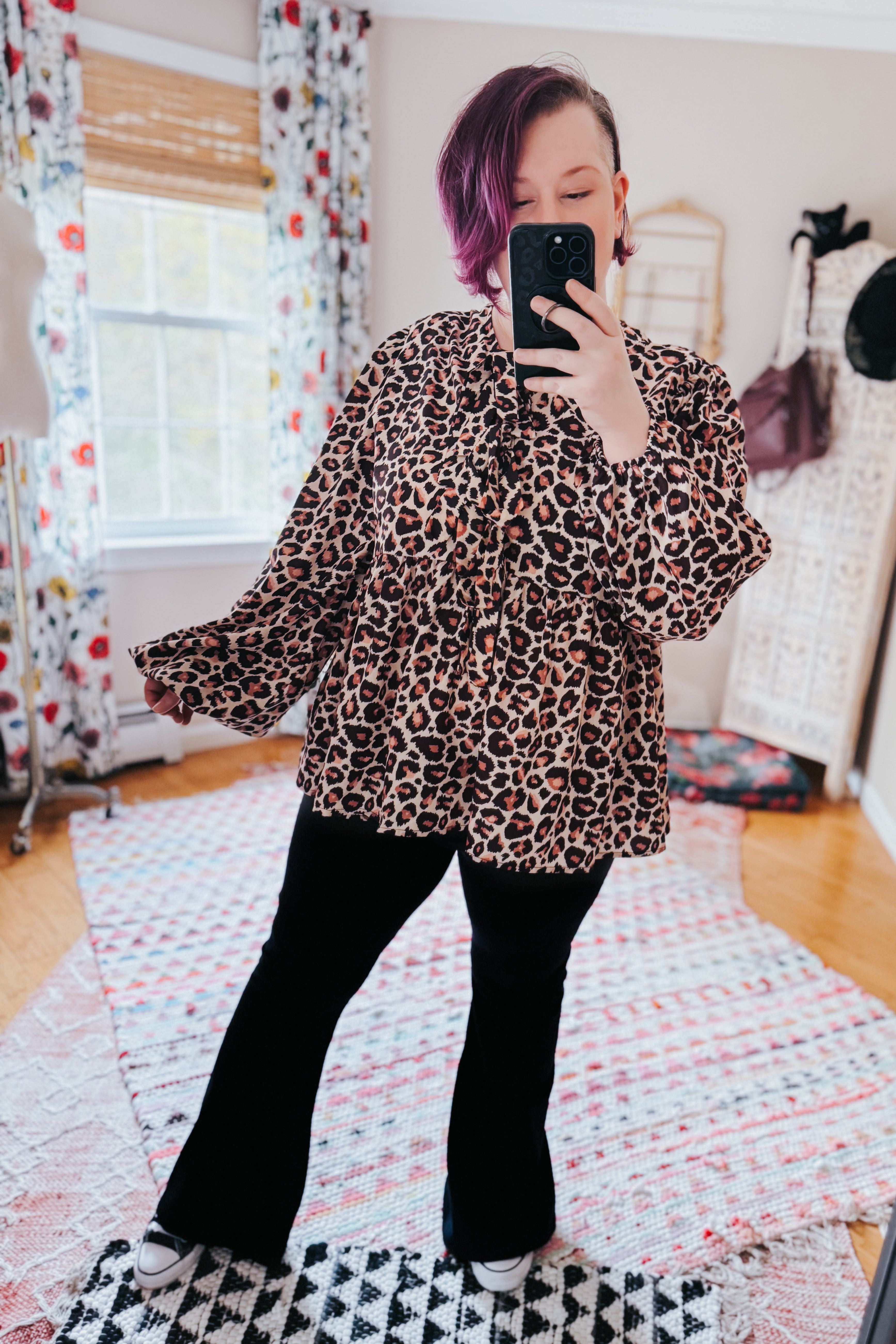 Woman taking mirror selfie wearing brown leopard print babydoll blouse with long sleeves paired with black pants in cozy room