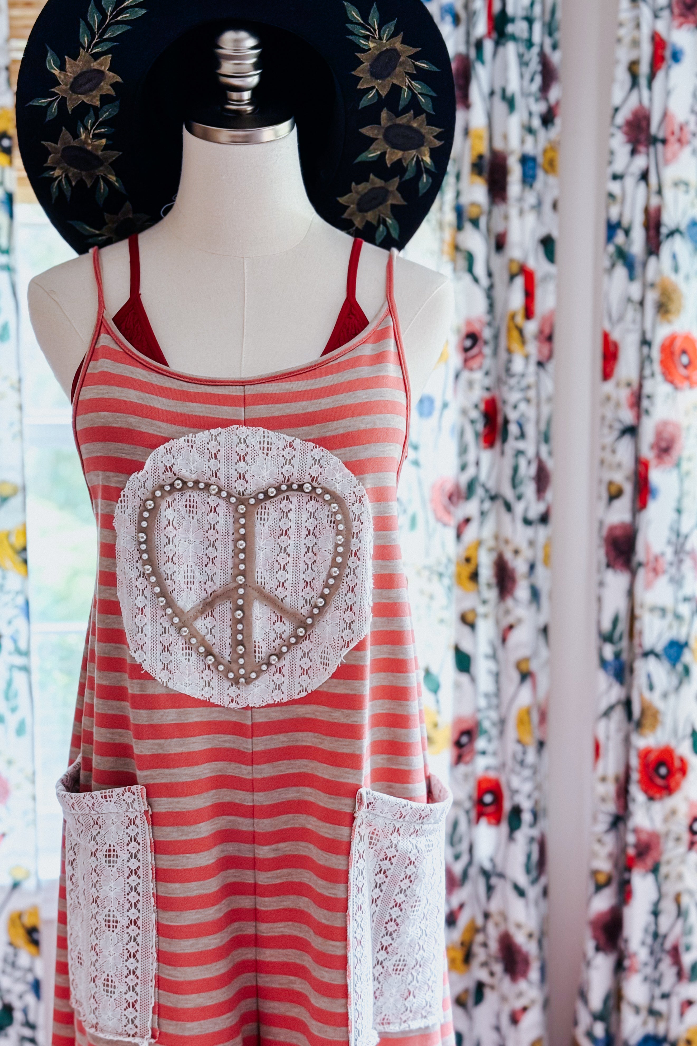 Striped red jumpsuit with lace-trimmed pockets and heart-shaped peace emblem displayed on mannequin with black floral hat