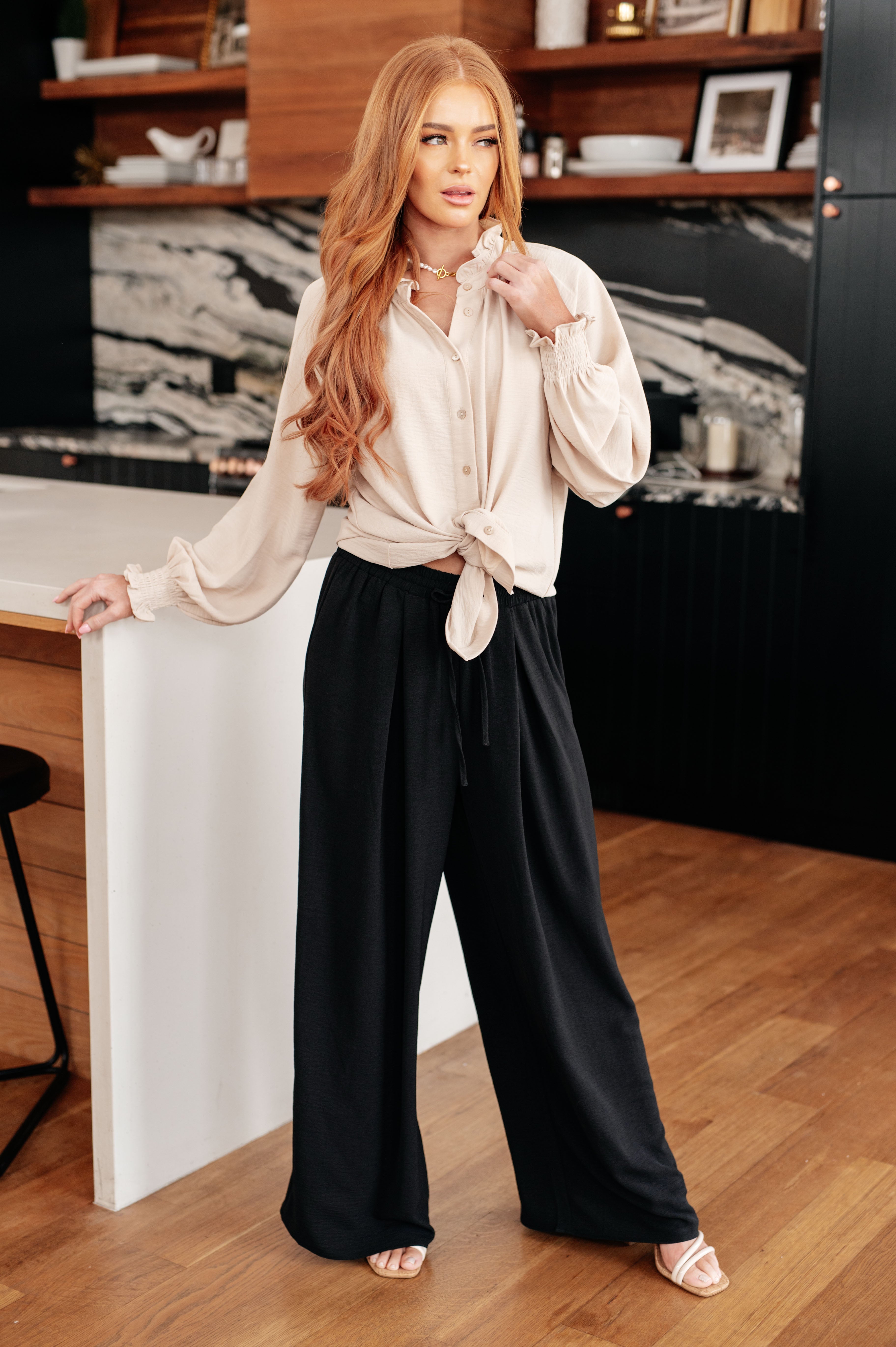 Woman wearing cream button-up blouse with tie front and voluminous sleeves paired with black wide-leg pants indoors
