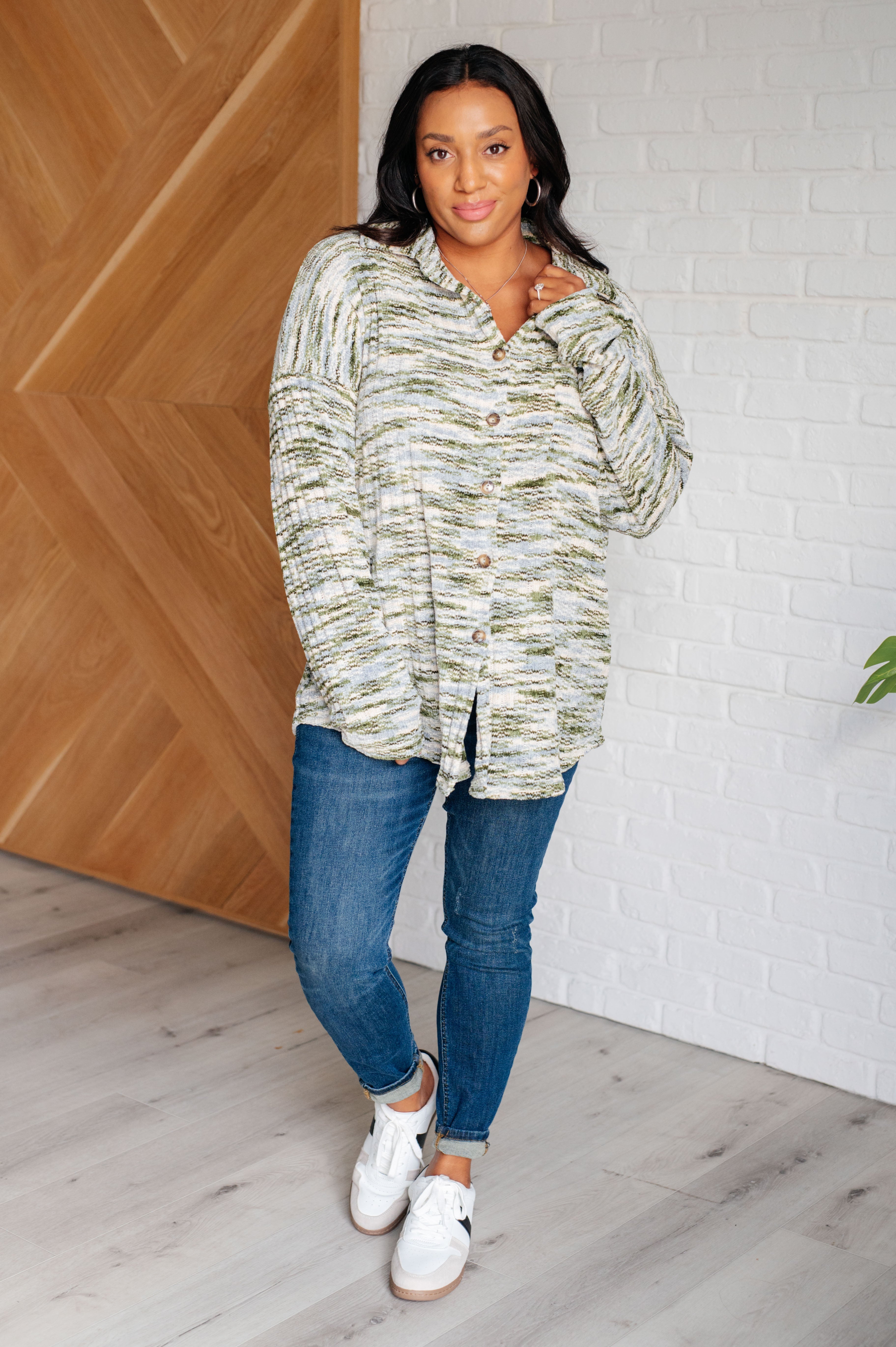 Woman wearing a green and cream knit shacket with jeans and white sneakers indoors