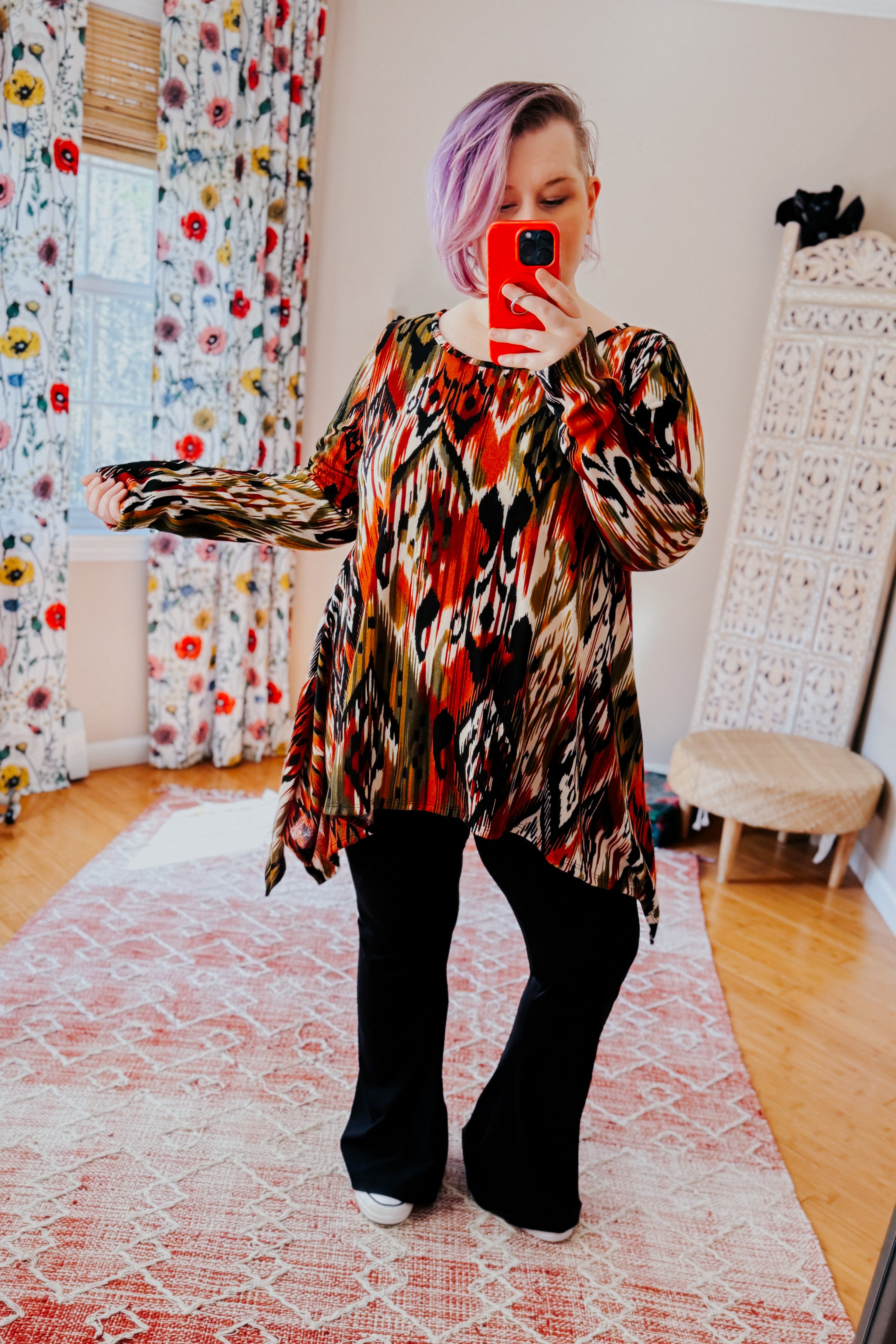 Woman with light purple hair wearing colorful abstract patterned long sleeve asymmetrical top and black pants indoors taking selfie
