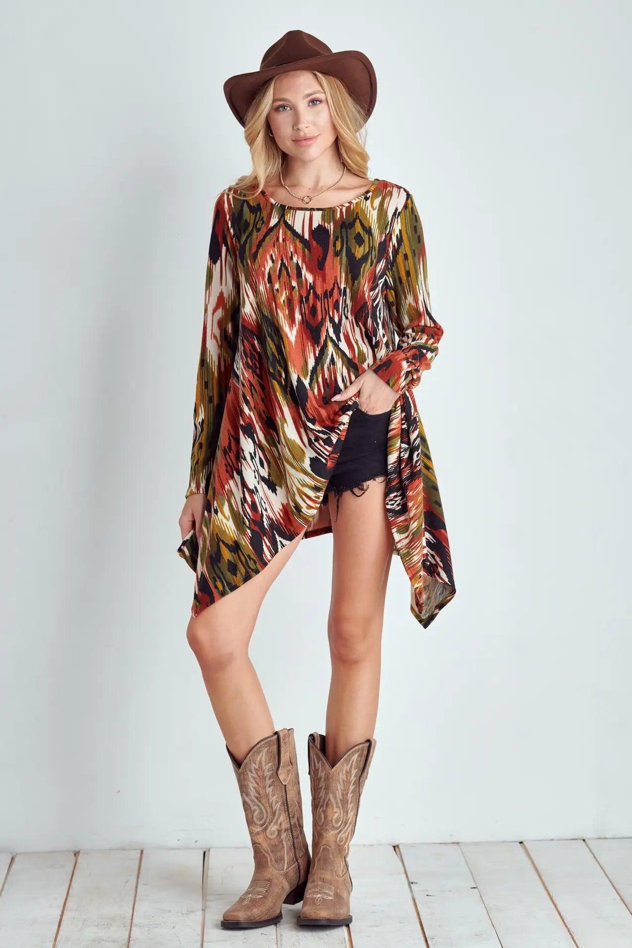 Woman wearing a long sleeve multicolor tribal patterned shark bite hem top with black shorts, cowboy boots, and brown hat