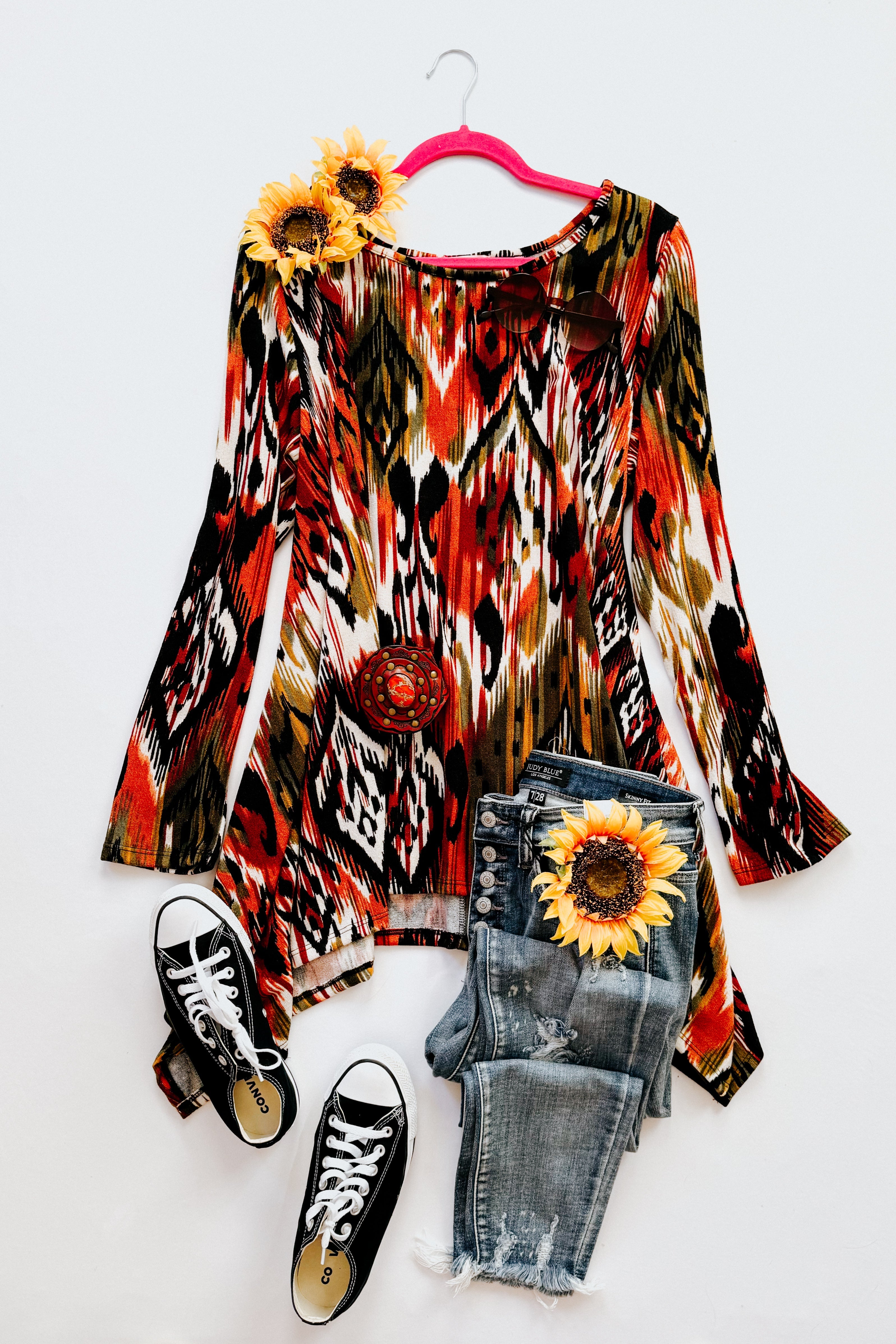 Red, green, and black geometric patterned long sleeve shark bite hem top displayed with jeans, sneakers, and sunflowers