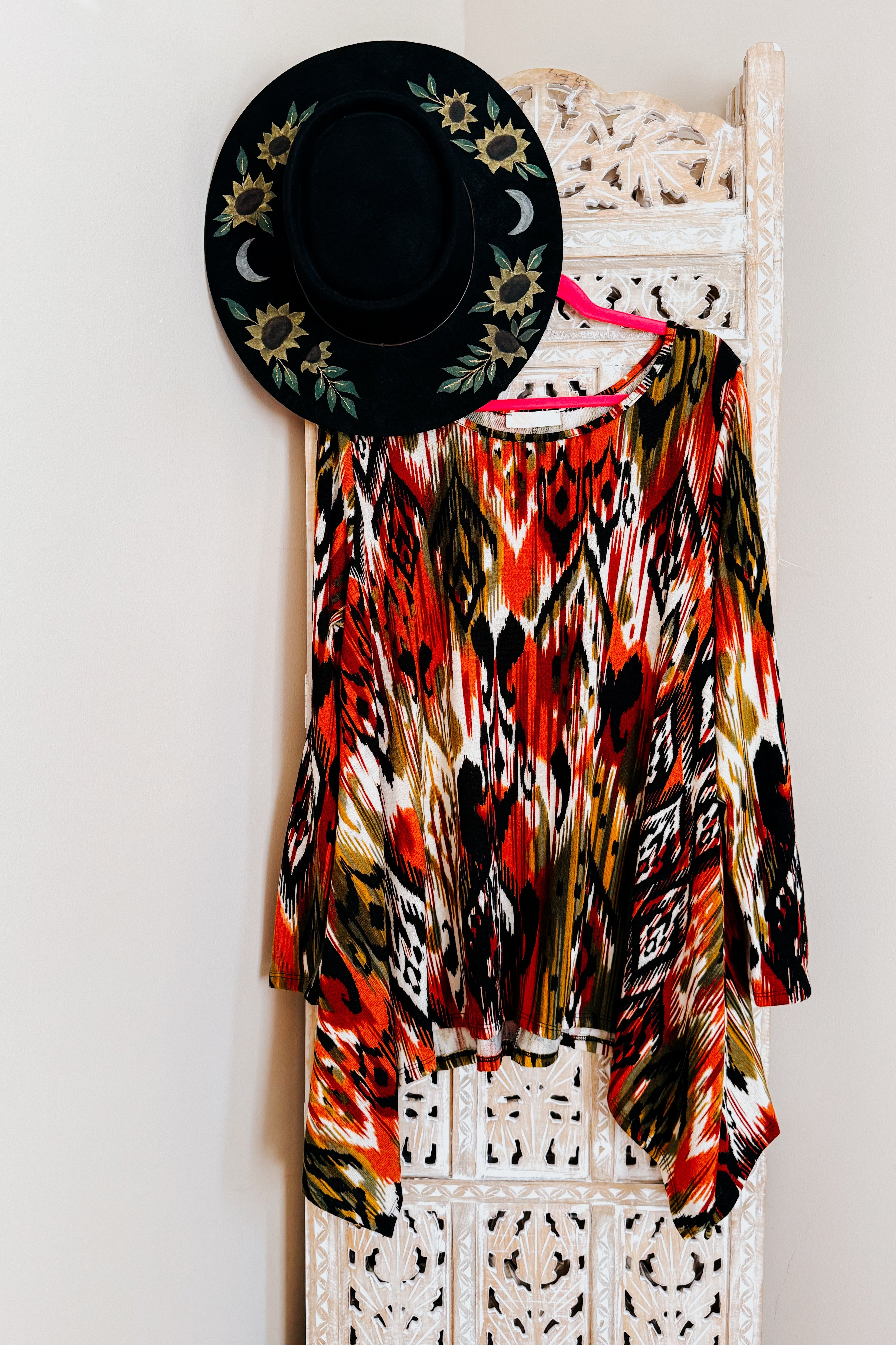 Long sleeve flowing red, green, and black geometric patterned shark bite hem top hanging on pink hanger with black floral hat