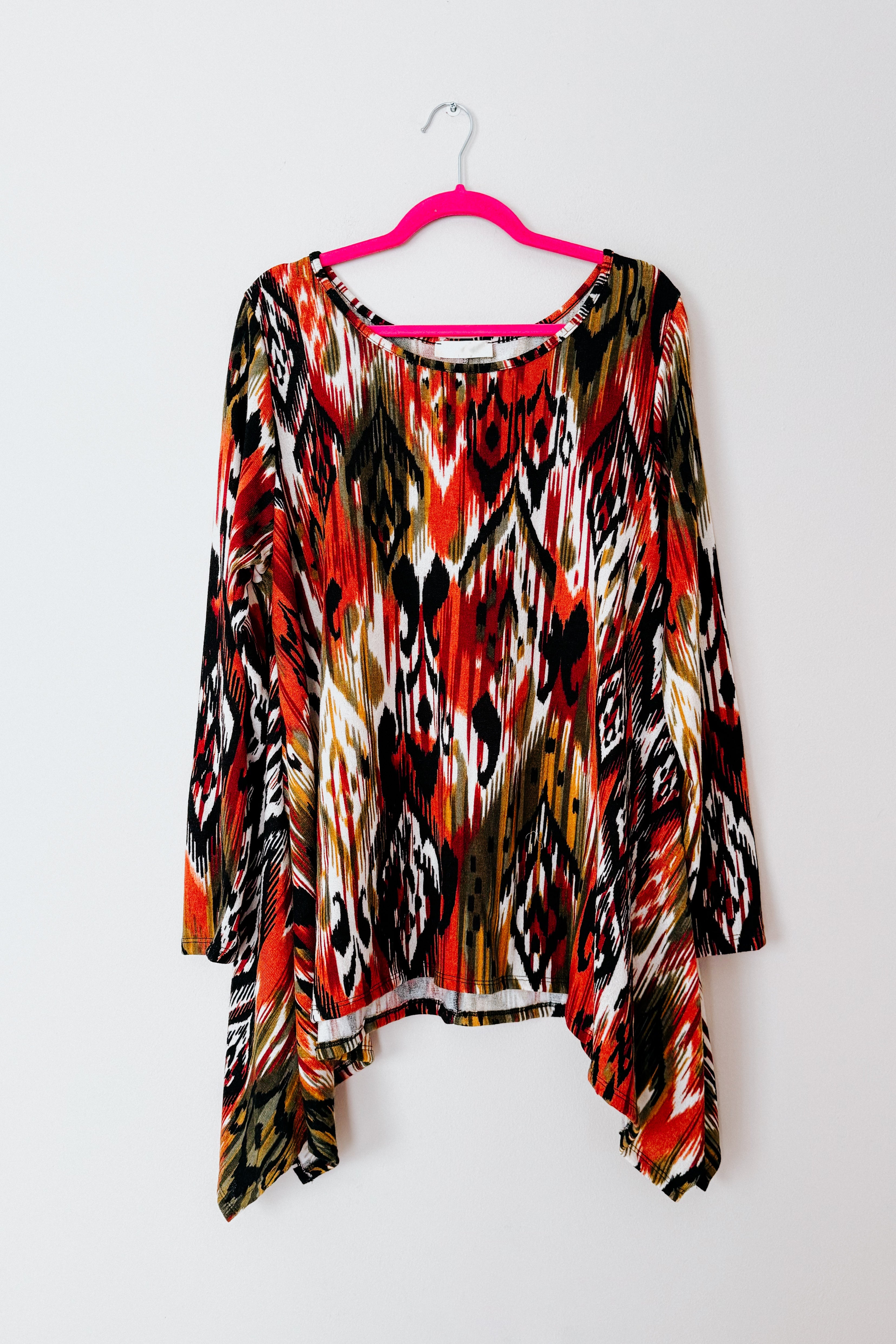 Colorful red, black, and olive patterned long sleeve shark bite hem top hanging on pink hanger against white backdrop
