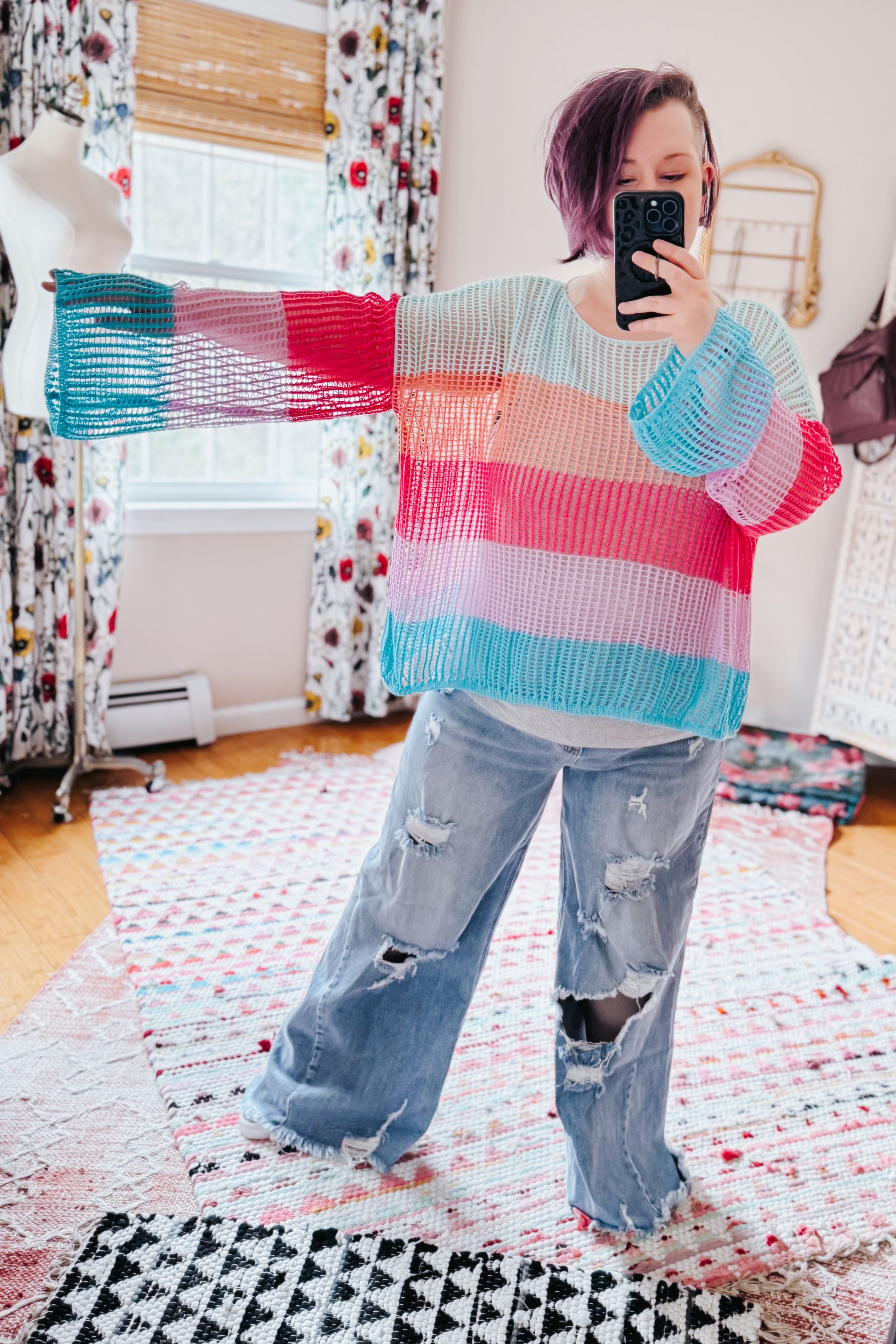 Woman taking mirror selfie wearing multicolor striped crochet mesh pullover with wide bell sleeves and distressed jeans