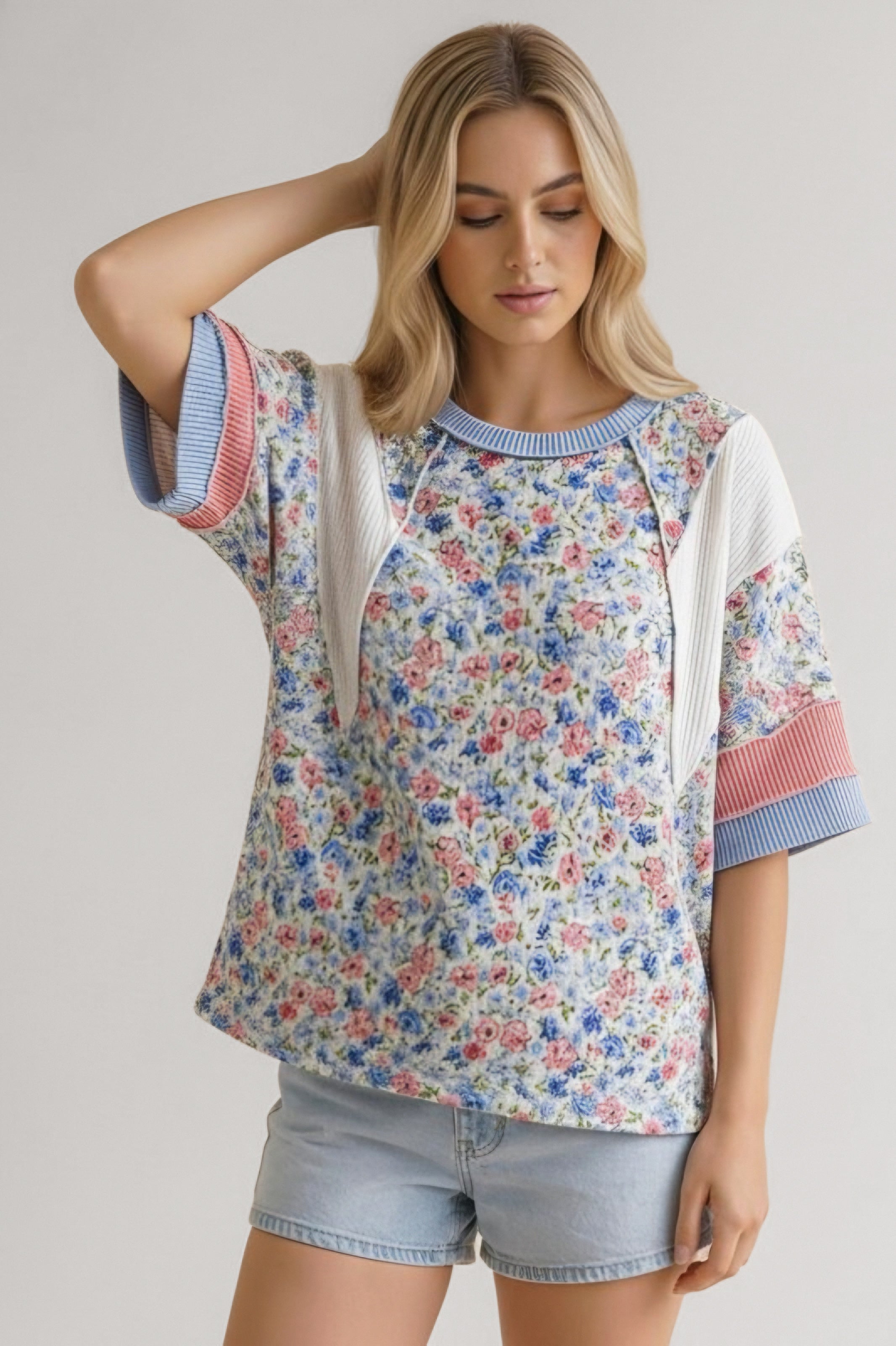 Woman wearing blue floral mixed media top with ribbed accents and light denim shorts standing against neutral background