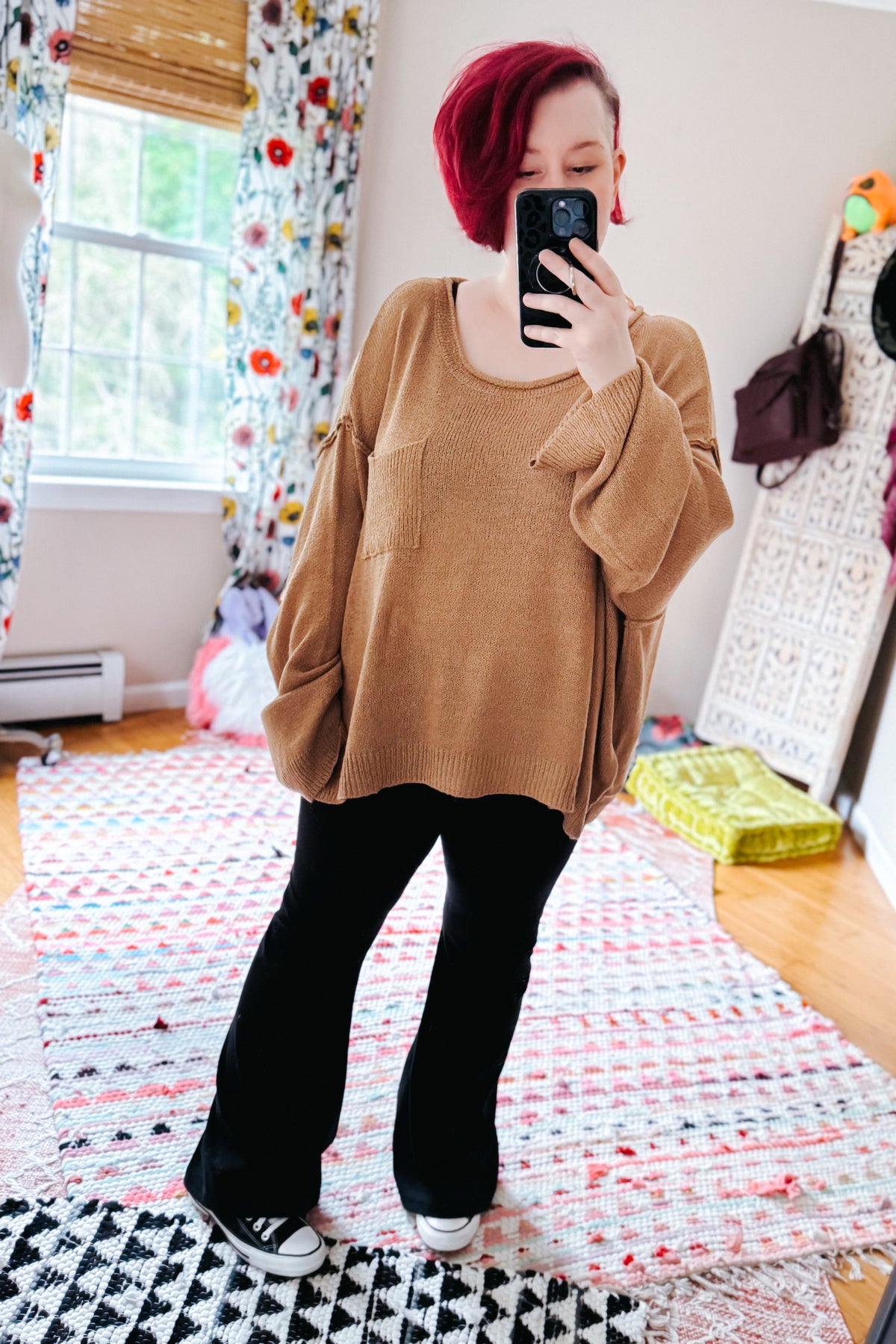 Red haired woman taking a bedroom mirror selfie in a camel boatneck sweater with chest pocket black pants and white sneakers on a colorful rug