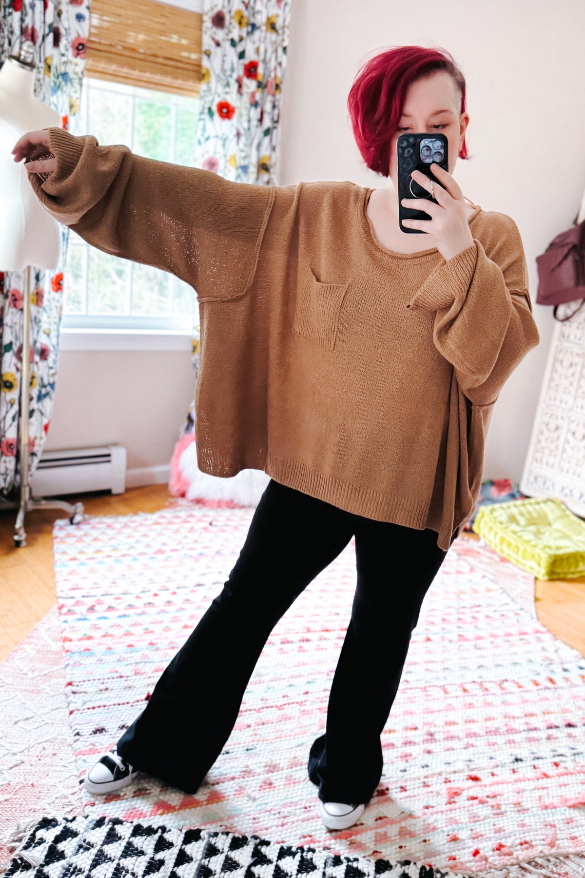 Red haired woman taking a bedroom mirror selfie in a camel boatneck sweater with dropped shoulders, black pants, and white sneakers