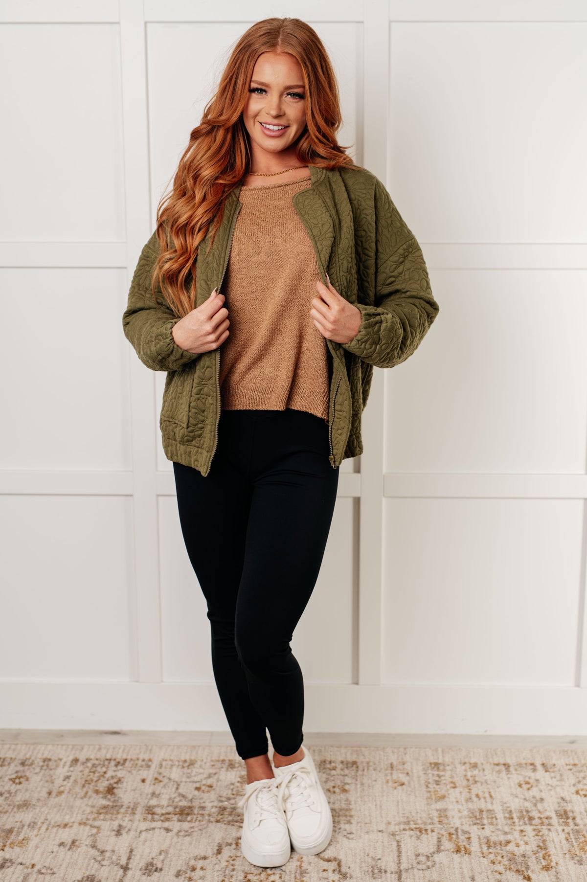 Model wearing a camel boatneck sweater with exposed seams beneath an olive cardigan styled with black pants and white sneakers