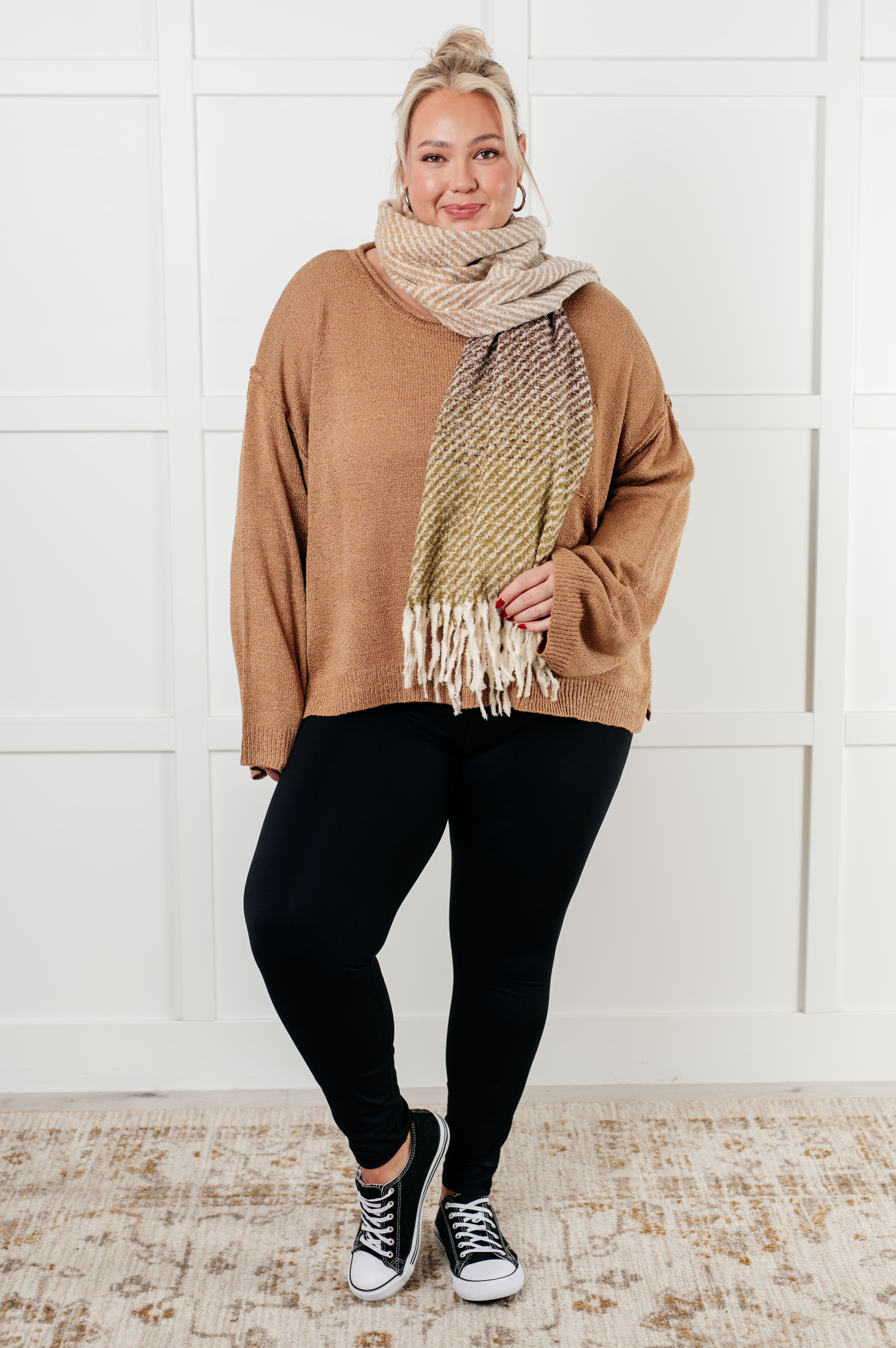 Plus size model wearing loose camel knit boatneck sweater with long sleeves, black leggings, striped fringed scarf, and black sneakers