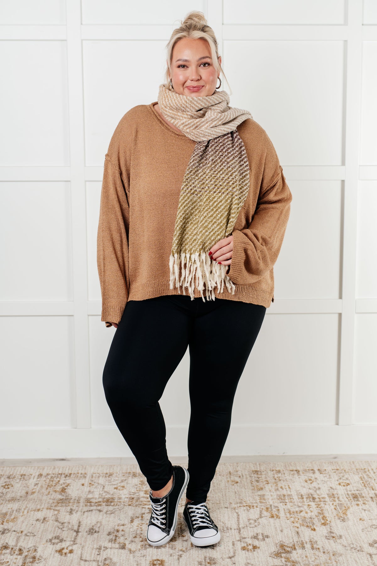 Model in a camel boatneck sweater with dropped shoulders styled with a fringed scarf black leggings and high top sneakers indoors
