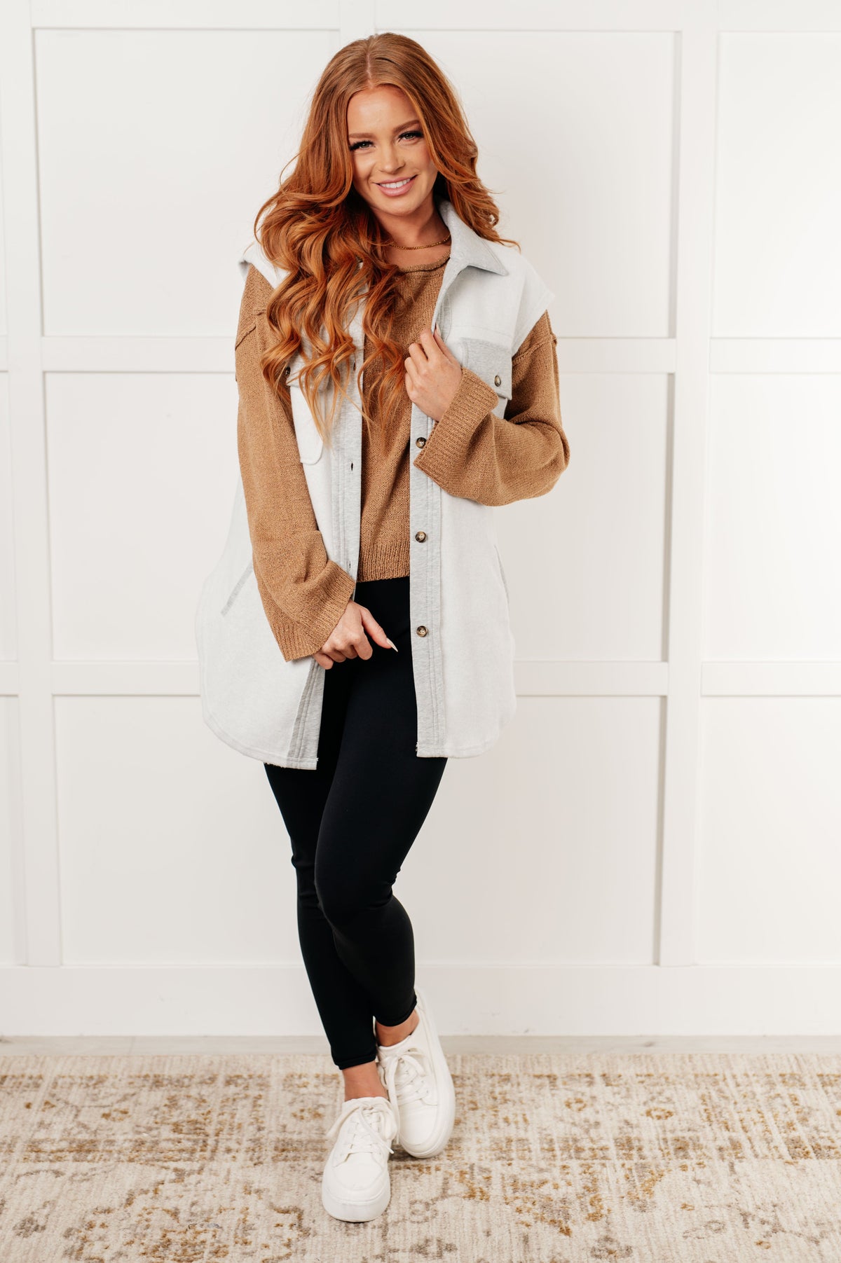Model wearing a camel boatneck sweater with a light vest, black leggings, and white sneakers in a bright paneled room