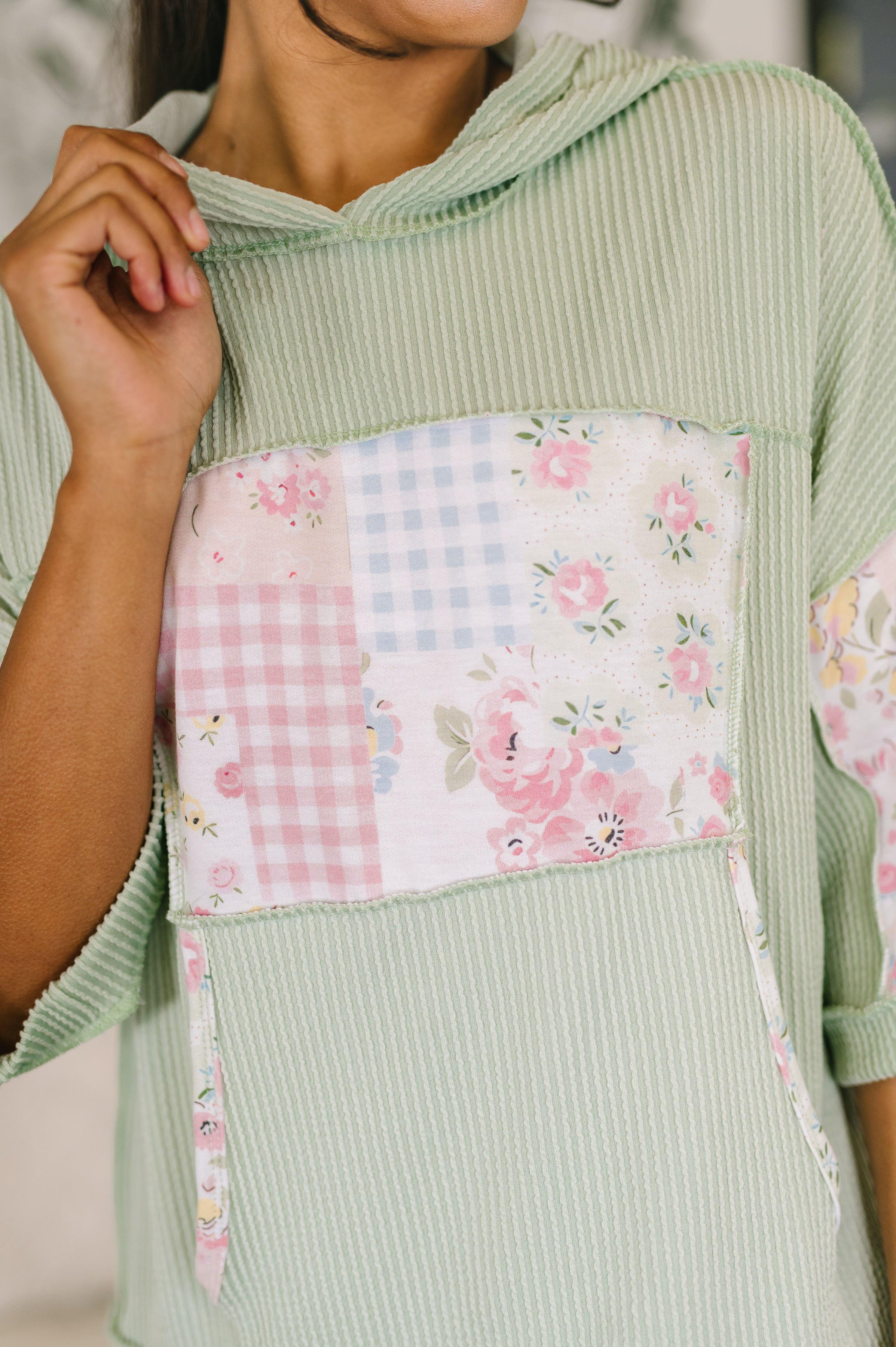 Close-up of light green ribbed knit hoodie with floral patchwork kangaroo pocket and half sleeves worn indoors