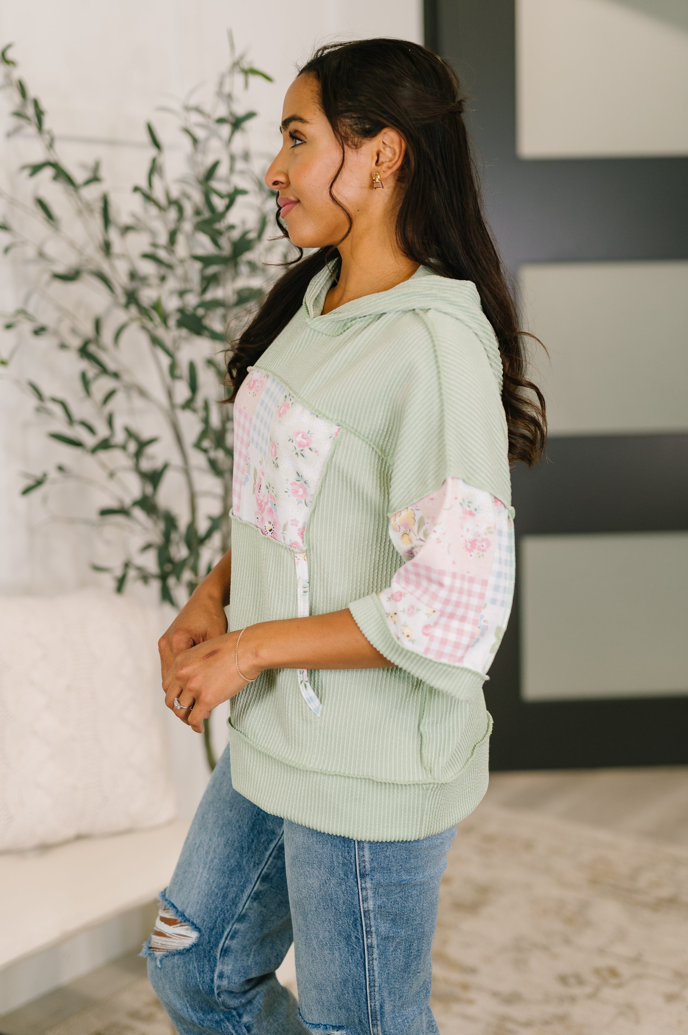 Side profile of woman wearing green ribbed knit hoodie with floral patchwork sleeves and distressed jeans indoors