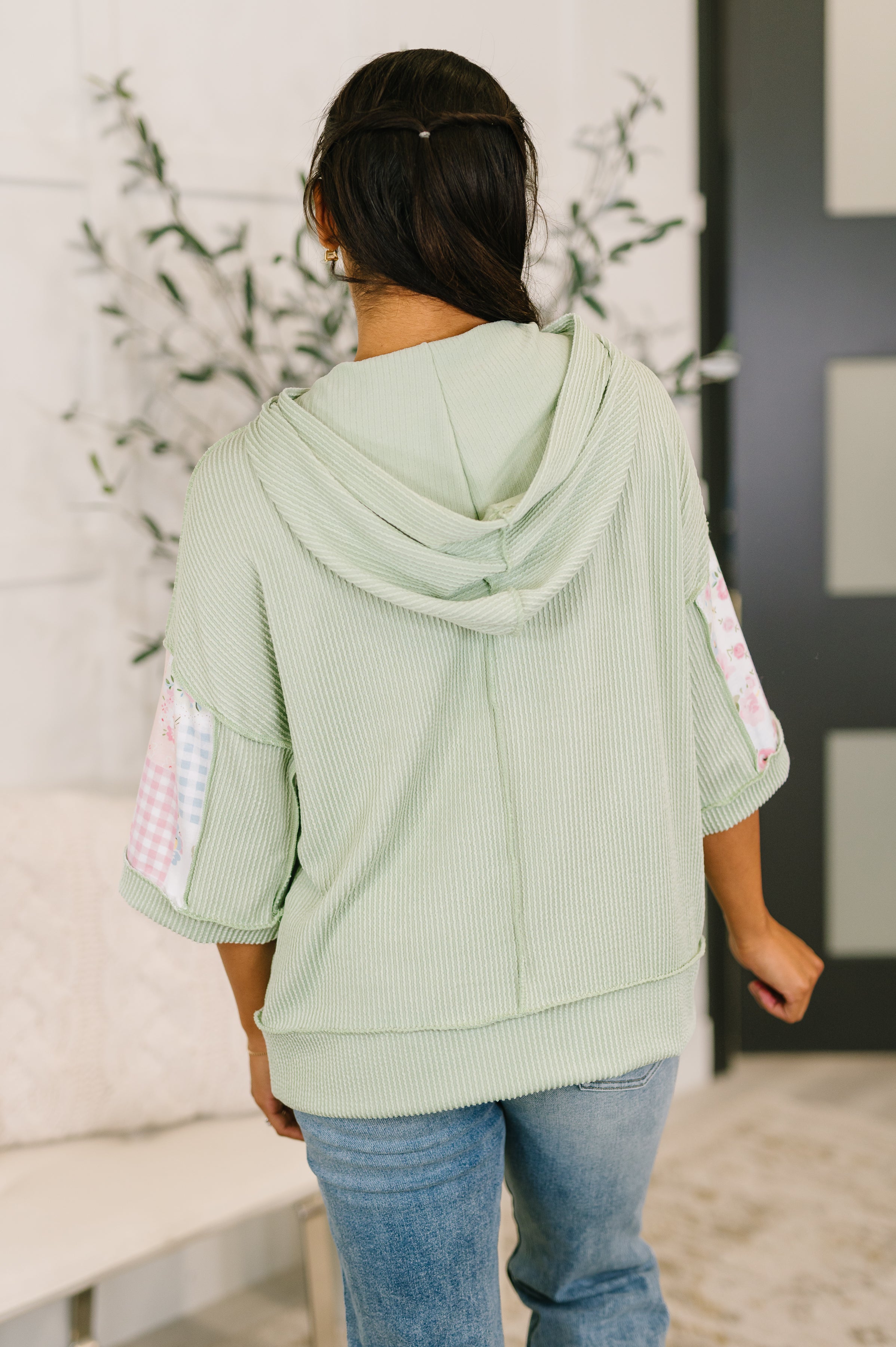 Back view of woman wearing light green cable knit half sleeve hooded top with patterned patchwork sleeves indoors